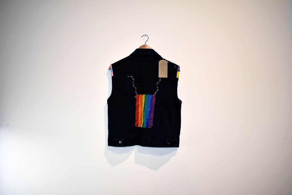 A dark vest with rainbow fabric hangs against a plain white wall.