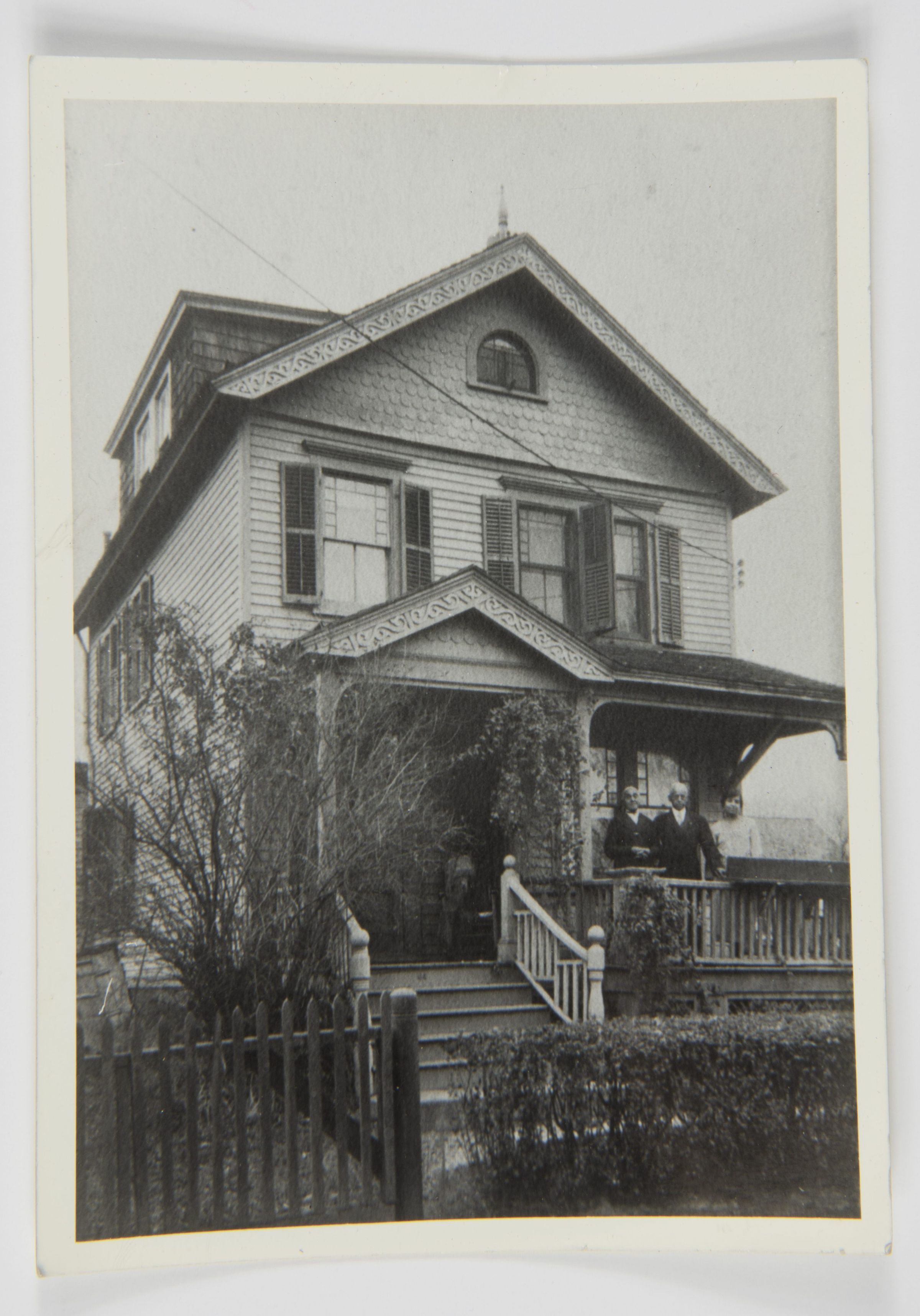 Latimer Home; two-story, features intricate detailing on its gables and three people standing on the porch; The black-and-white photograph depicts a vintage two-story house with architectural details that include ornate trim along the gable roof and a dormer window. The facade features wood siding and shutters, with several of the windows open, allowing a glimpse of the interior. Climbing vines and bushes adorn the exterior, adding a hint of nature to the scene. A small staircase with a decorative railing leads up to the covered porch, where three individuals stand, creating a sense of human presence and perhaps indicating a family dwelling. The wooden picket fence encircles the front yard, which is landscaped with trimmed hedges, suggesting care and maintenance. The overall atmosphere conveys a sense of nostalgia and timelessness, evoking memories of a bygone era. The details within the image capture the charm and character of traditional residential architecture, showcasing the craftsmanship and style of the period.
