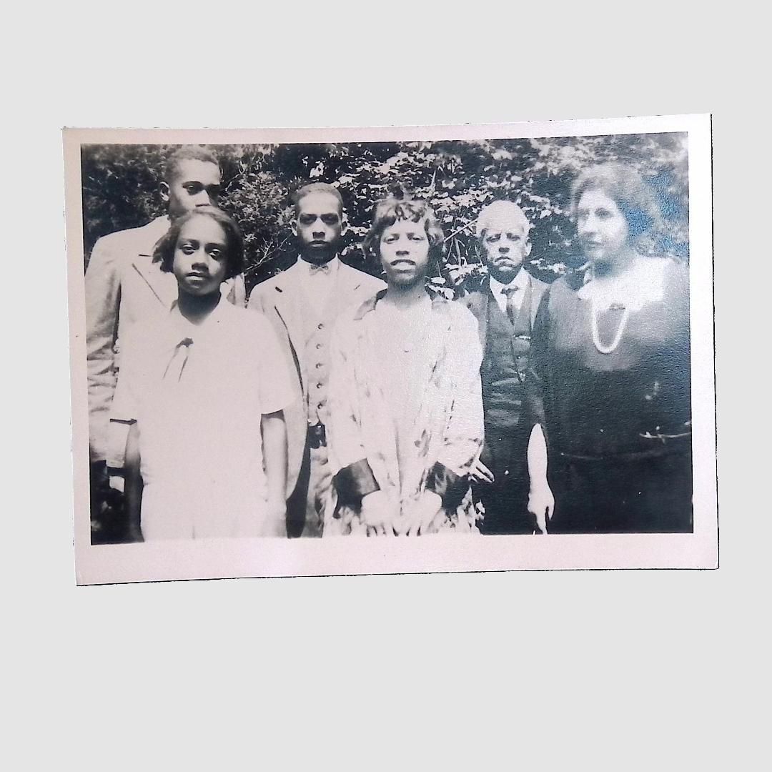 A group of six Latimer family members pose outdoors in black-and-white vintage attire, surrounded by greenery.; The image is a black and white photograph depicting six individuals, comprising both men and women, in formal attire, standing together and posing for the camera. Three men are dressed in suits and ties, with one man in the back row holding a serious expression. The women are wearing dresses, one of which is adorned with a necklace. The background features foliage, suggesting an outdoor setting, and the individuals appear to be in a garden or park-like area. The image conveys a sense of formality and appears to be from an earlier 20th-century period, capturing a moment of significance or celebration for the group.