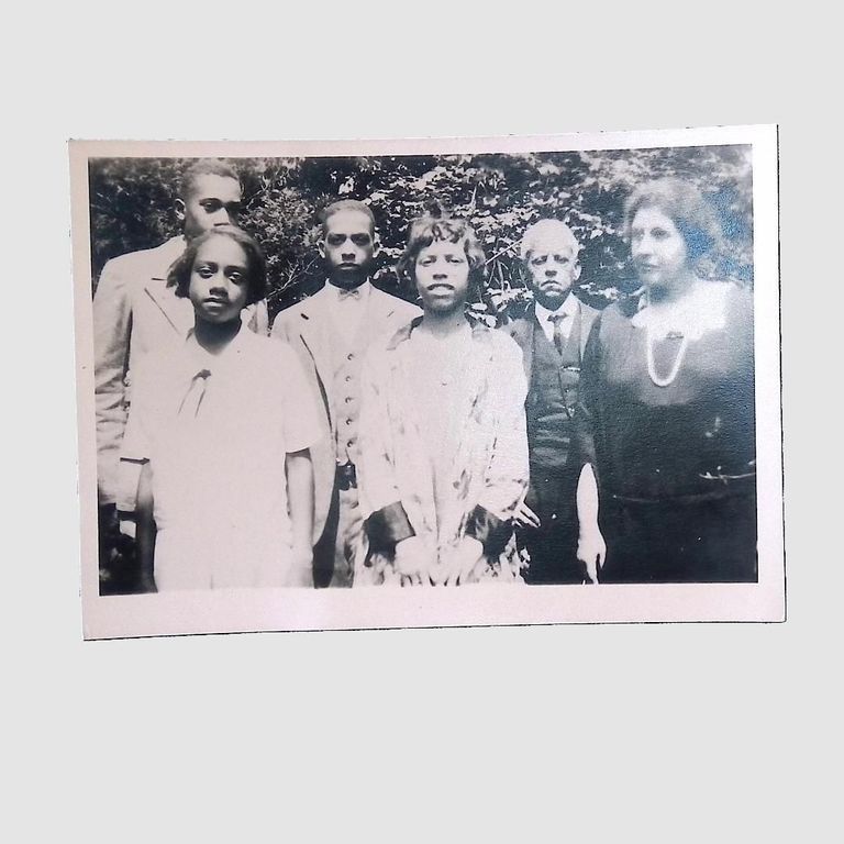 A group of six Latimer family members pose outdoors in black-and-white vintage attire, surrounded by greenery.; The image is a black and white photograph depicting six individuals, comprising both men and women, in formal attire, standing together and posing for the camera. Three men are dressed in suits and ties, with one man in the back row holding a serious expression. The women are wearing dresses, one of which is adorned with a necklace. The background features foliage, suggesting an outdoor setting, and the individuals appear to be in a garden or park-like area. The image conveys a sense of formality and appears to be from an earlier 20th-century period, capturing a moment of significance or celebration for the group.