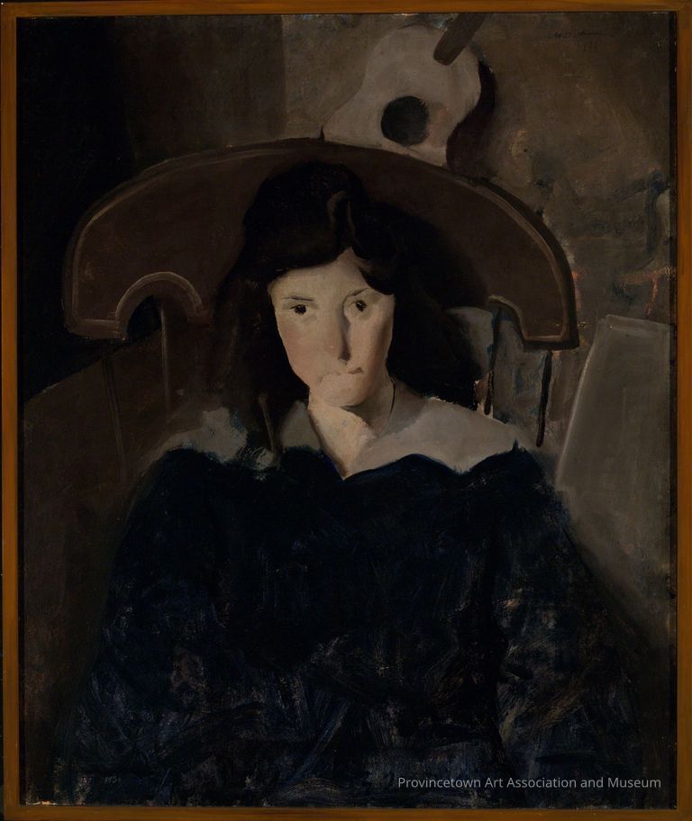 Portrait of young woman with dark hair and blue dress, sitting in chair, dark interior with guitar behind her