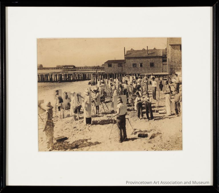 Students crowd the beach for a Hawthorne painting class. Equipped with easels and canvases, the students paint figures and portraits. To block the glaring sun, straw hats adorn the tops of heads, while feet dig into the beach sand. In the background of the photograph, piers line the shoreline, standing tall from the shallows of low tide sand.