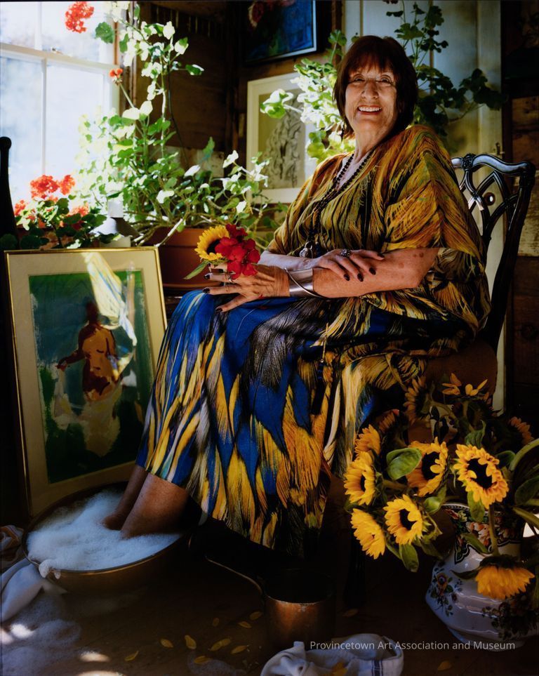Photo of gallerist Berta Walker sitting with her feet in a soapy bowl, in a colorful dress and surrounded by art and flowers
