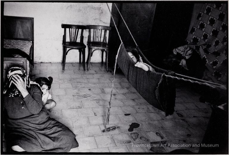 Interior; woman covering her face sitting on the floor with a child behind her; child sleeping in hammock
