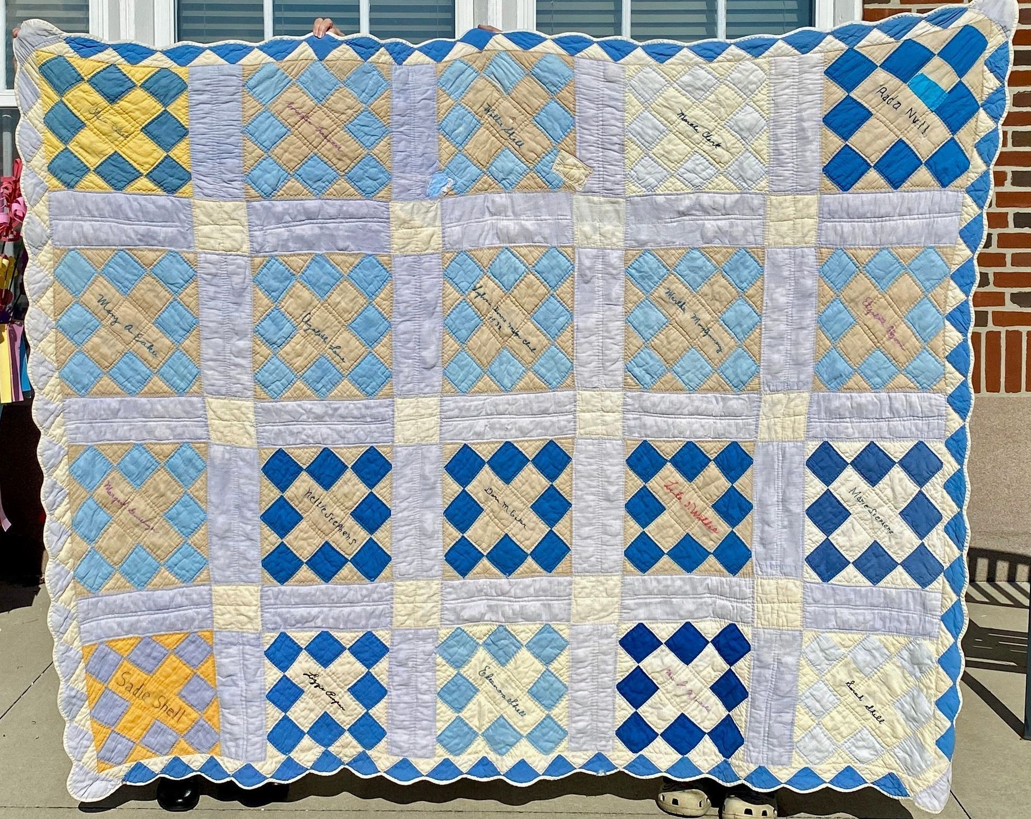 Sylvan Home Makers Club 1932 signature quilt: Photo taken in front of the former Will Mayfield College Arts and Sciences Building.
The quilt has a pattern of alternating squares in shades of blue, cream, and yellow, bordered with a scalloped edge; each square is stitched with various names, representing contributions from different individuals. These quilts were a collaborative or commemorative project that reflects a community or special event.