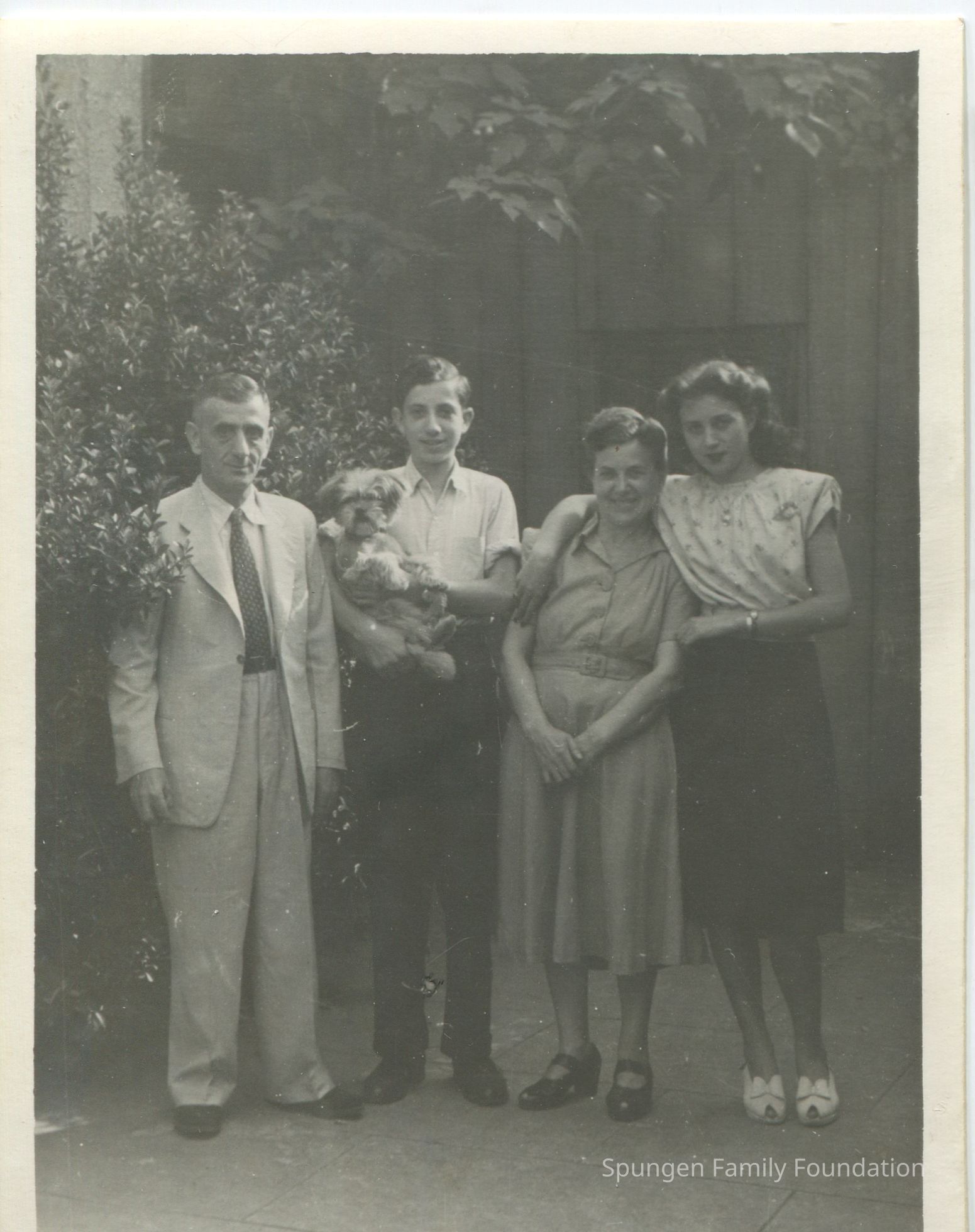 c.1940s-1950s, Front side - Photograph of the Sternberg Family.: Black-and-white photograph depicting the Sternberg Family. At the left, Herman, is wearing a grey or beige suit with dark shoes and a pattern tie. He has a short dark haircut. To the right, Gary, is holding a dog in his arms. He is wearing a white collard shirt, black pants, and black shoes. He has a short dark haircut. To the right, Augusta, is wearing a neutral colored dress with black wedge heels. She has her dark hair in an up-do. She is holding her hands together in front. To the right, Ruth, is wearing a pattern short sleeve blouse with a black skirt and white heels. She has a long dark haircut. Her right hand is over Augusta's shoulder. The family is posed in front of a residential building. Green foliage as a bush and tree leaves are present in the background.