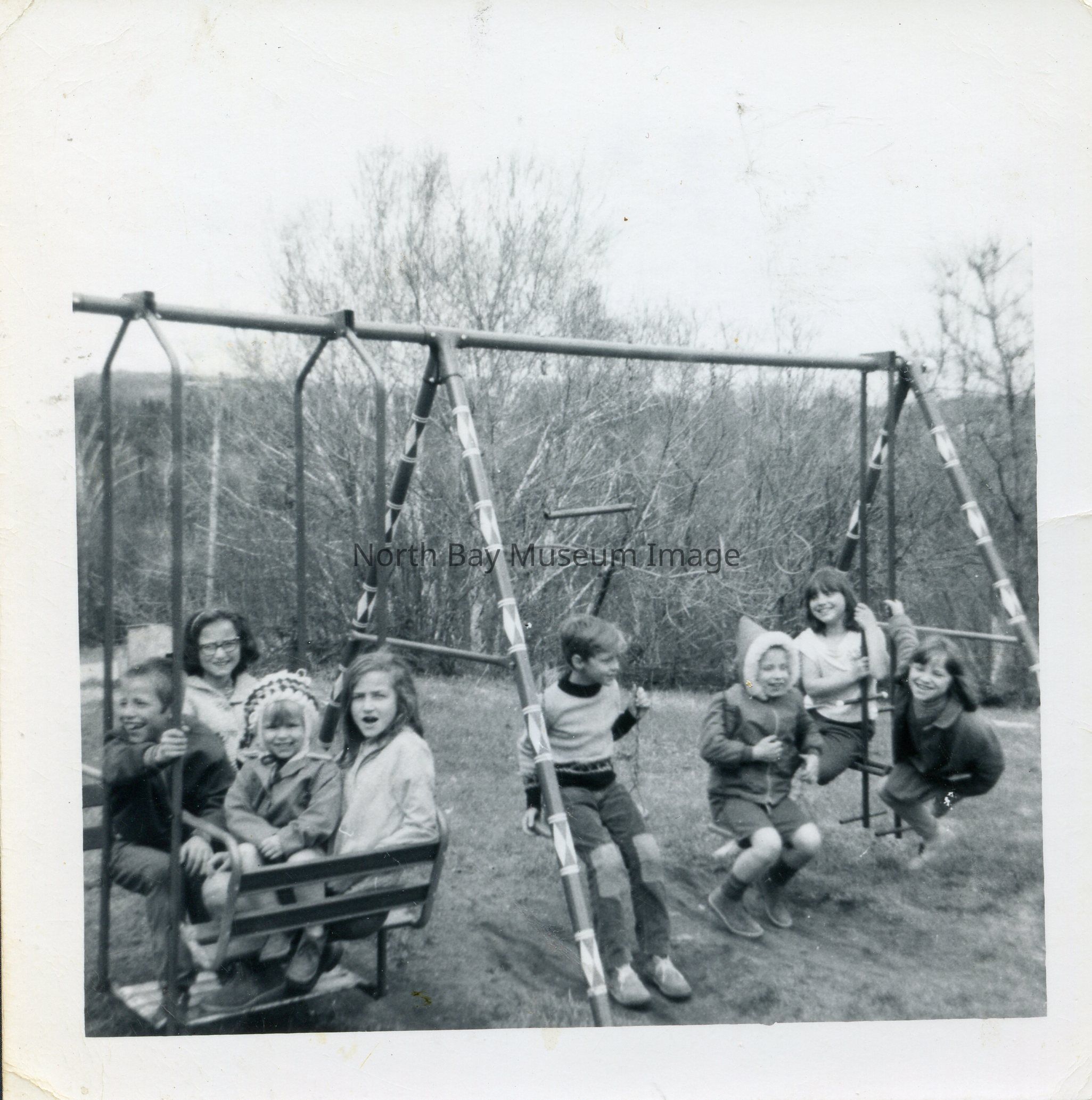 2025.01.018: A whole flock of children mill about the swing-set, likely at Mrs Rousseau's residence. At least one member of the Triplets is present at the far left, possibly Garry. Her four older children seem to be spread among the swings with three girls (friends or relatives of the family). It seems to be the early Spring or late Fall due to the attire and lack of tree leaves.