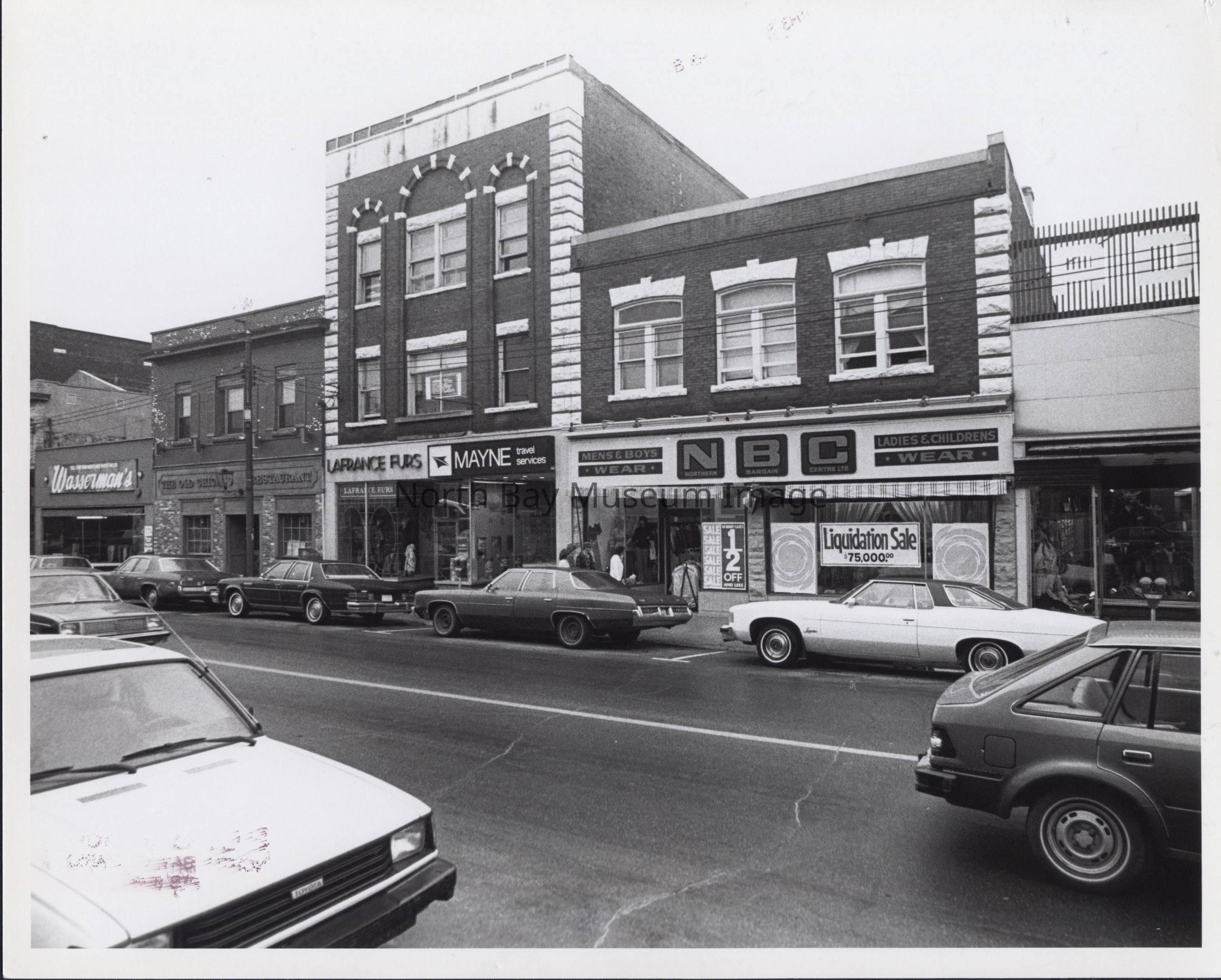 2025.04.005: Main Street, on the opposite side of the Mayne Travel Services building from 2025.04.004. From left, Wasserman's (?), the Old Chicago Cafe, LaFrance Furs, Mayne Travel Services, and Northern Bargain Centre Ltd. (which seems to be liquidating). The inscription suggests it was made by the PL Dept, which may be the police dept(?).