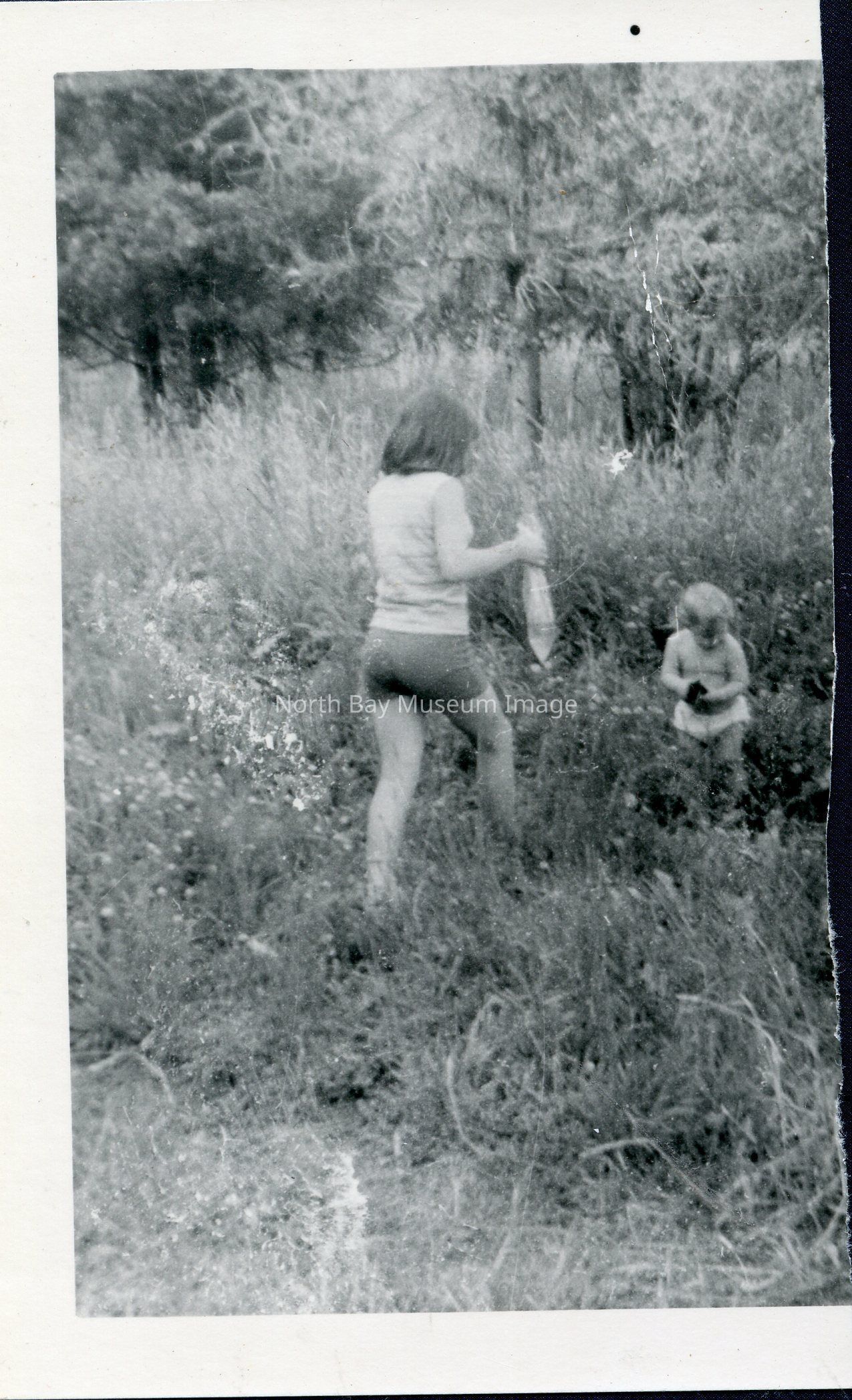 2025.01.022: Jane (or a family friend or relative) and a Rousseau Triplet run amok among a heath. The former holds aloft a bag with an oblong shape therein, possibly a bottle. The tot holds a small dark object. Likely part of the same picnic series of photos in 2025.01.011 and 2025.01.014, this photo has been cut in half.