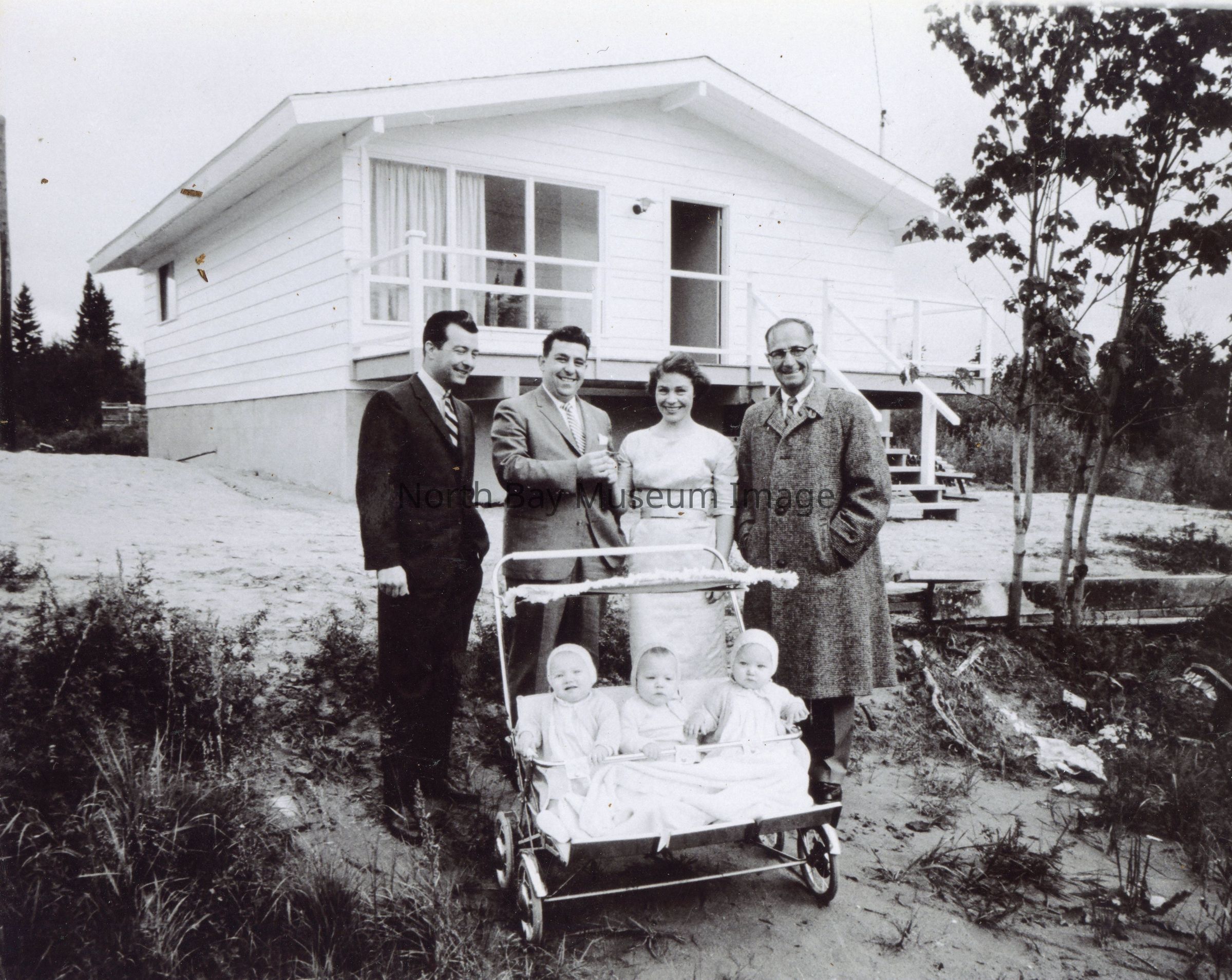 2025.01.030: This Nugget photo is part of the report on Birdie receiving the keys to her new house built on the initiative of the Jaycees, Friday 15 September 1961. To commemorate the contributions of the community, an open-house was held Sunday 17 Sept, before the family moved in Monday 18 Sept, in time to celebrate the first birthday of the Triplets Tuesday 19 September. From left, Charles M Scott, Jaycee member and former employer of Wilfred Rousseau, John Palangio Jr, former president of the North Bay Jaycees, Birdie Rousseau, and Reeve George Coombes of West Ferris. Below sit Corrine, Garry, and Geraldine.