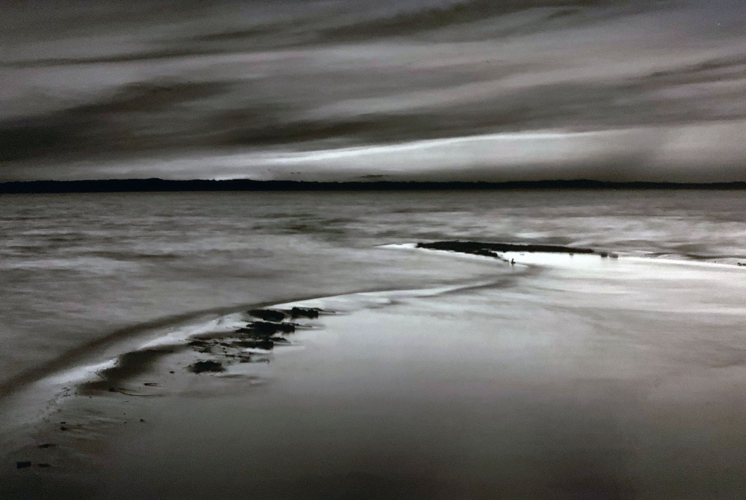 The black and white image captures a serene coastal landscape, where gentle waves lap against the shore under a dramatic cloudy sky, creating a tranquil and atmospheric scene. Shadows and highlights play across the sand and water, enhancing the peaceful yet mysterious ambiance, suggesting an early morning or late evening setting.