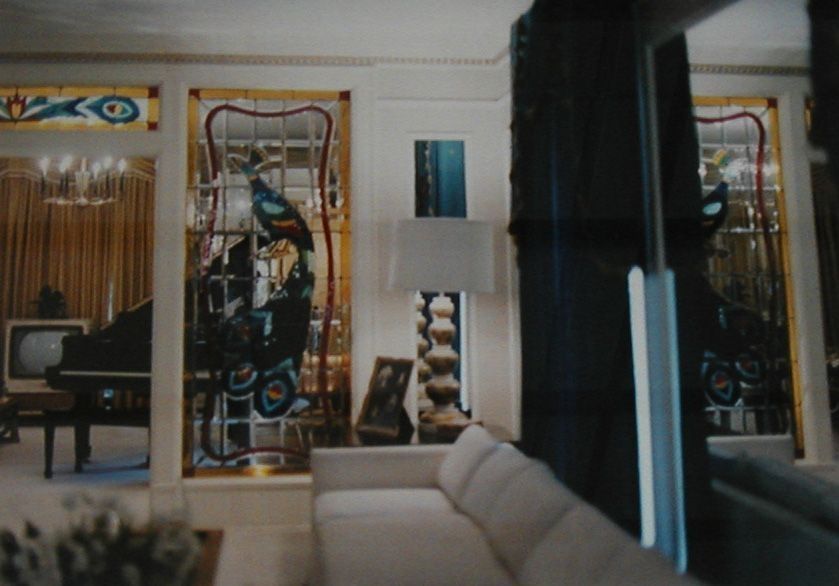 The image depicts a luxurious living room featuring an elegant white couch and a stylish standing lamp. The room is adorned with stained glass panels showcasing intricate peacock designs, adding a touch of sophistication and color. In the background, a grand piano rests beside an old-fashioned television, while a chandelier hangs from the ceiling.