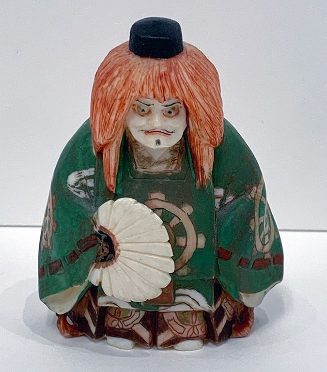 The image depicts a figurine representing a Noh theatre character, showcasing intricate details such as a vivid green kimono adorned with traditional patterns. The figure features orange, textured hair cascading down, accentuating their dramatic facial expression. Holding a delicate white fan, the figurine conveys a sense of poised elegance.