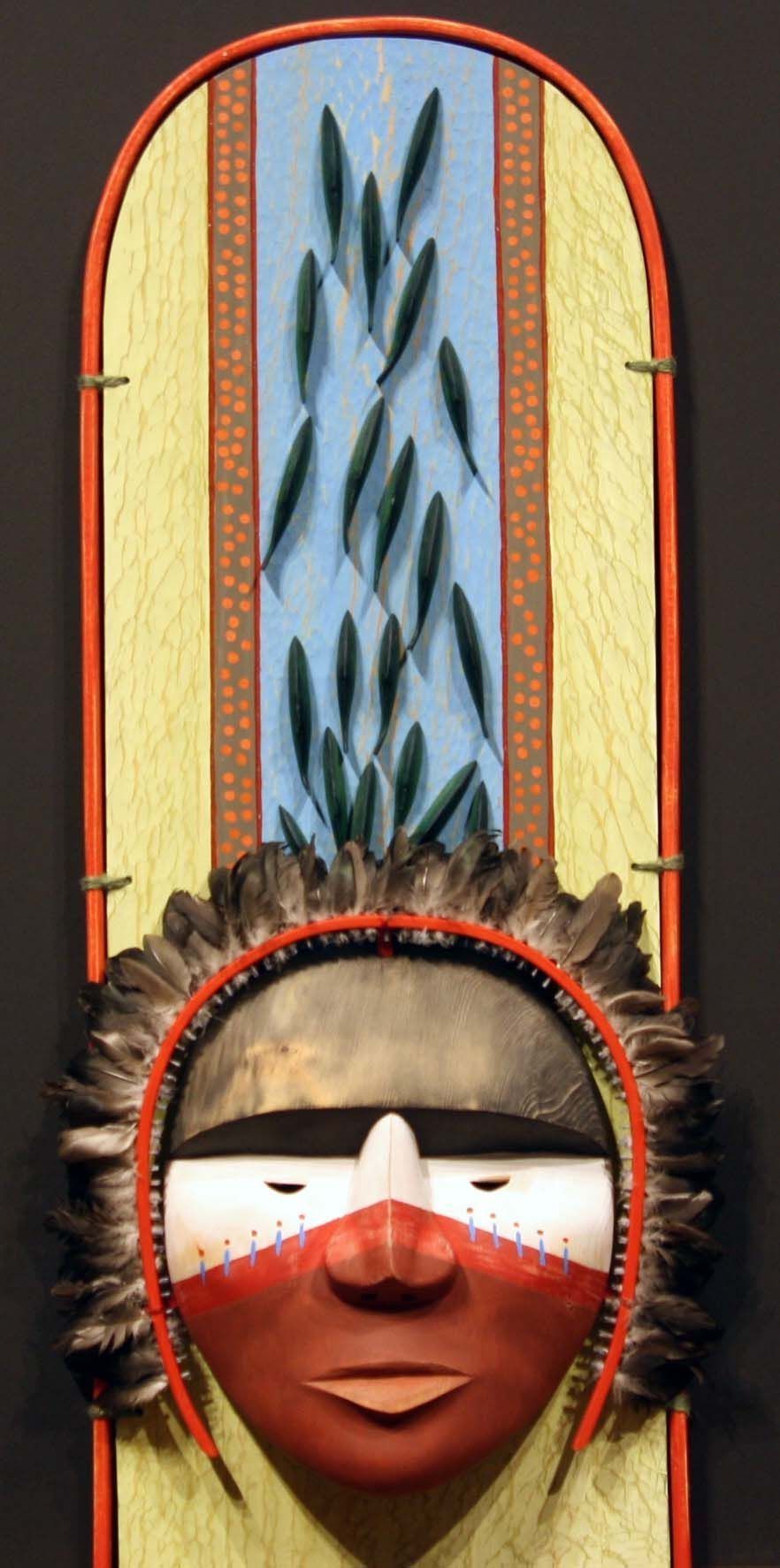 The image features an Alaskan mask with striking colors and patterns, prominently centered against a backdrop suggestive of salmon swimming upstream, evidenced by elongated, fish-like shapes in motion on a light blue panel. The mask itself has a fierce yet serene expression, with red and white painted details, and is adorned with feathers around the head, framed by a vertical structure with orange dot patterns on the sides.