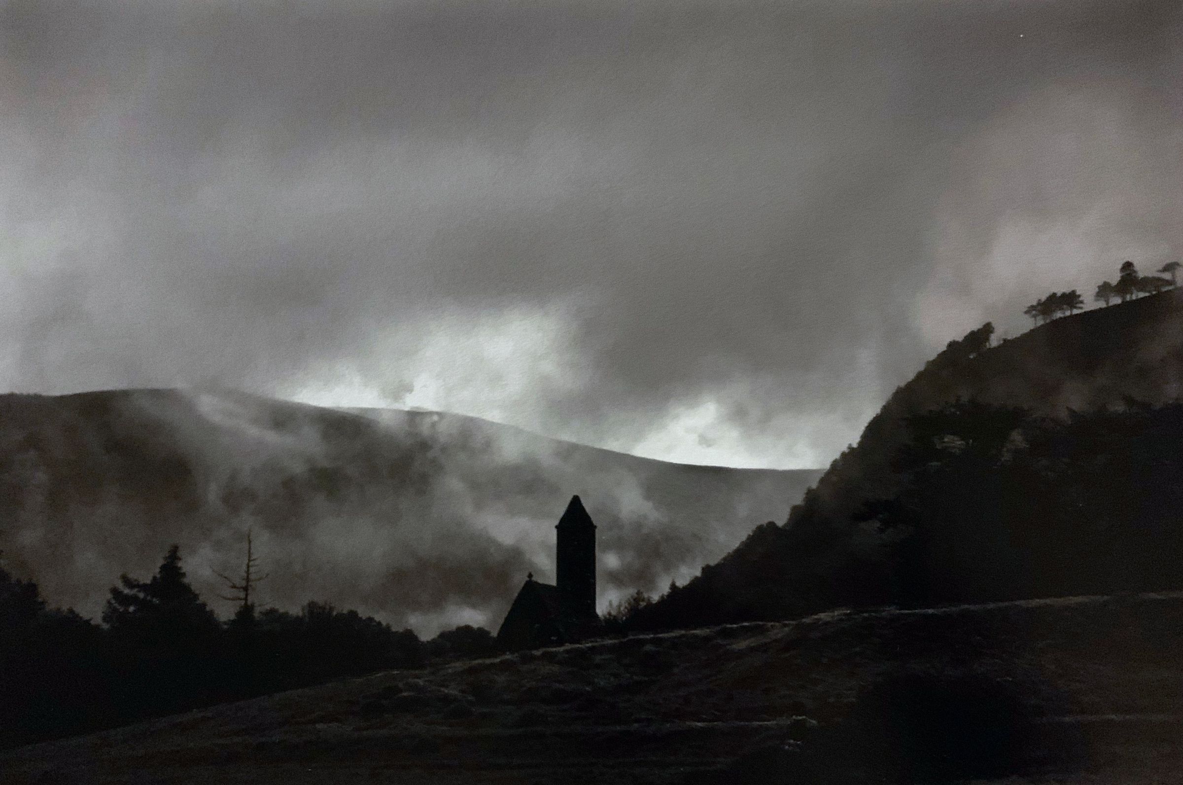In the haunting black and white image, a silhouette of a church tower emerges against misty hills and dense forests, all enveloped in atmospheric fog and low-hanging clouds, creating an eerie yet captivating landscape that evokes a sense of mystery and solitude, inviting the viewer's imagination to delve into the scene.
