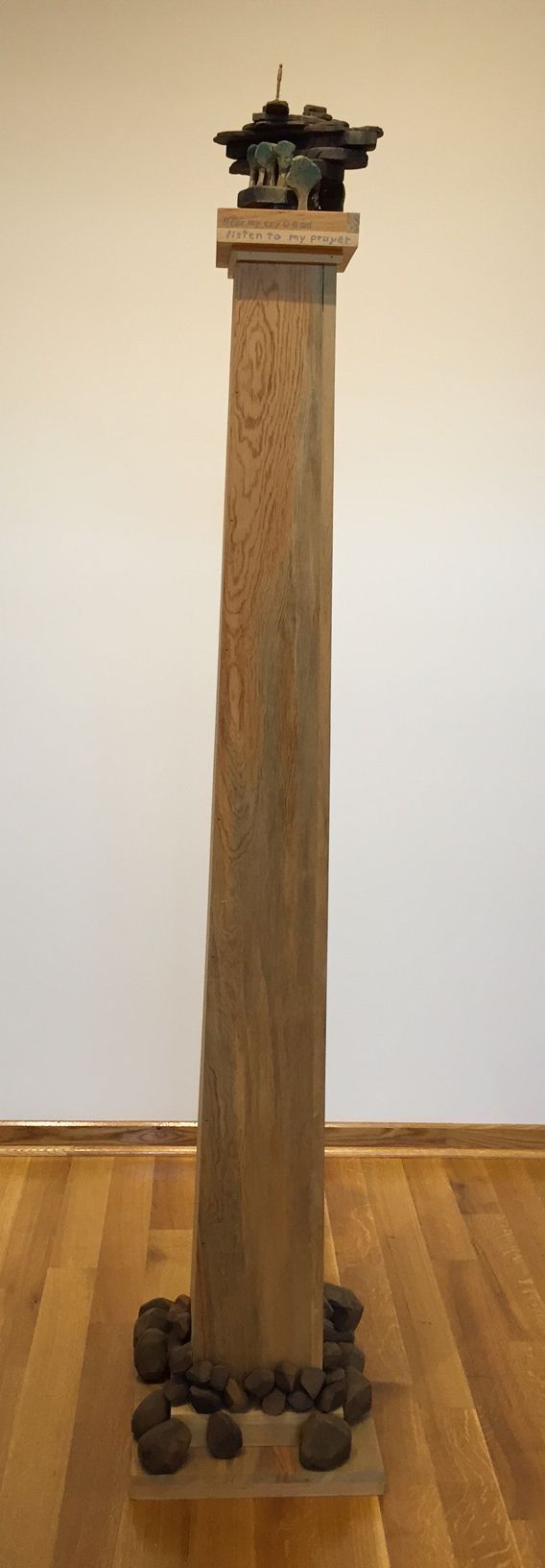 The image depicts a wooden sculpture featuring a small human figure standing atop stacked slabs, resembling rock formations, with miniature tree-like structures below, all set within a wooden frame that bears the inscription, "Hear my cry O God, listen to my prayer," creating a contemplative and spiritual composition.