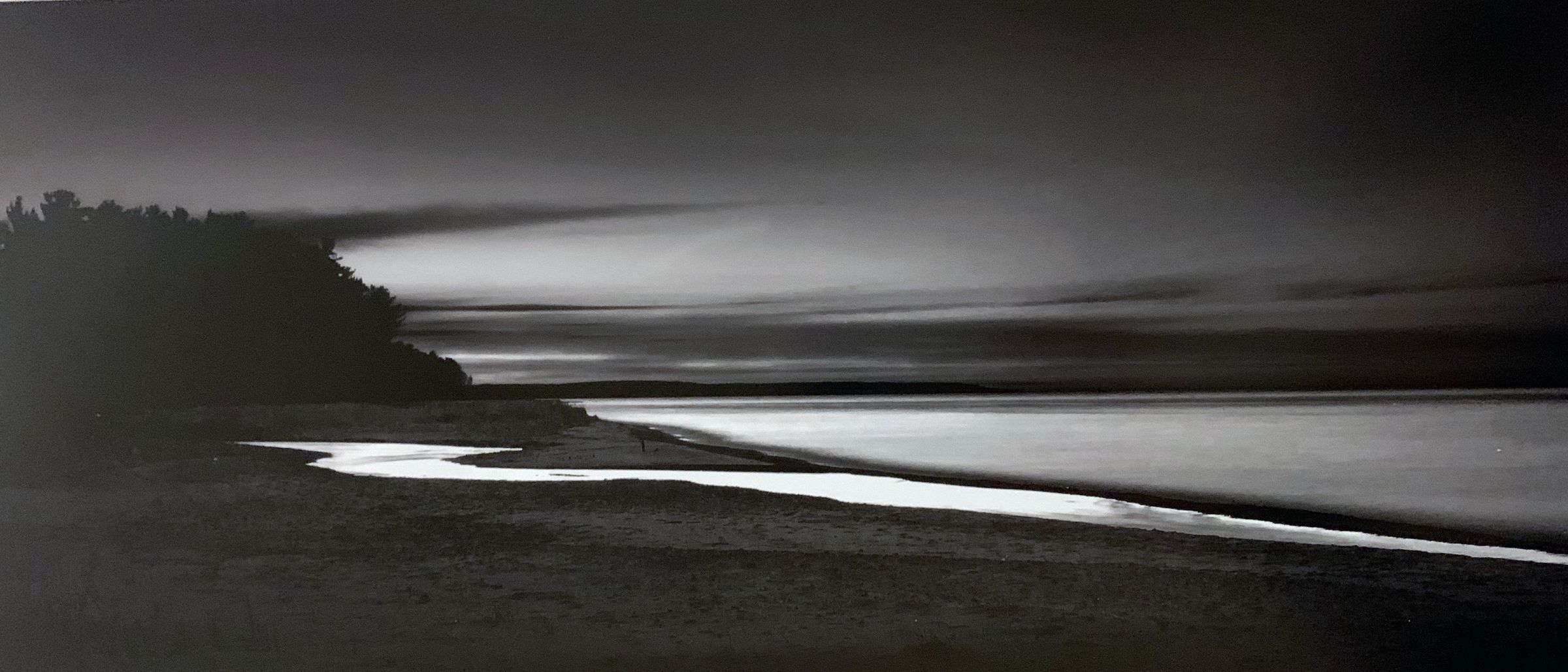 The black and white photograph captures a serene shoreline at dusk, with a meandering stream reflecting the fading light as it gently flows into the vast, calm sea beneath a dramatically clouded sky. A dense line of silhouetted trees stand quietly to the left, emphasizing the tranquil and contemplative mood of the photograph.