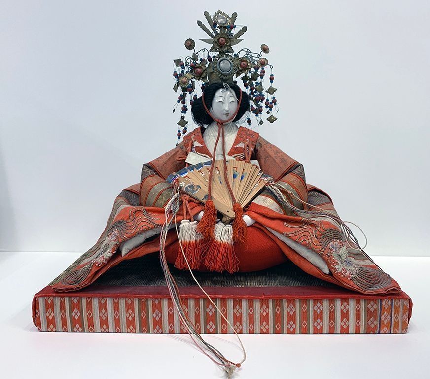 The image showcases an intricately designed traditional Japanese doll, adorned with an elaborate headdress and sitting elegantly on a platform. The doll wears a beautiful, ornate kimono with vibrant red and orange patterns, holding a delicate fan with tassels. The headdress features floral and ornamental details.