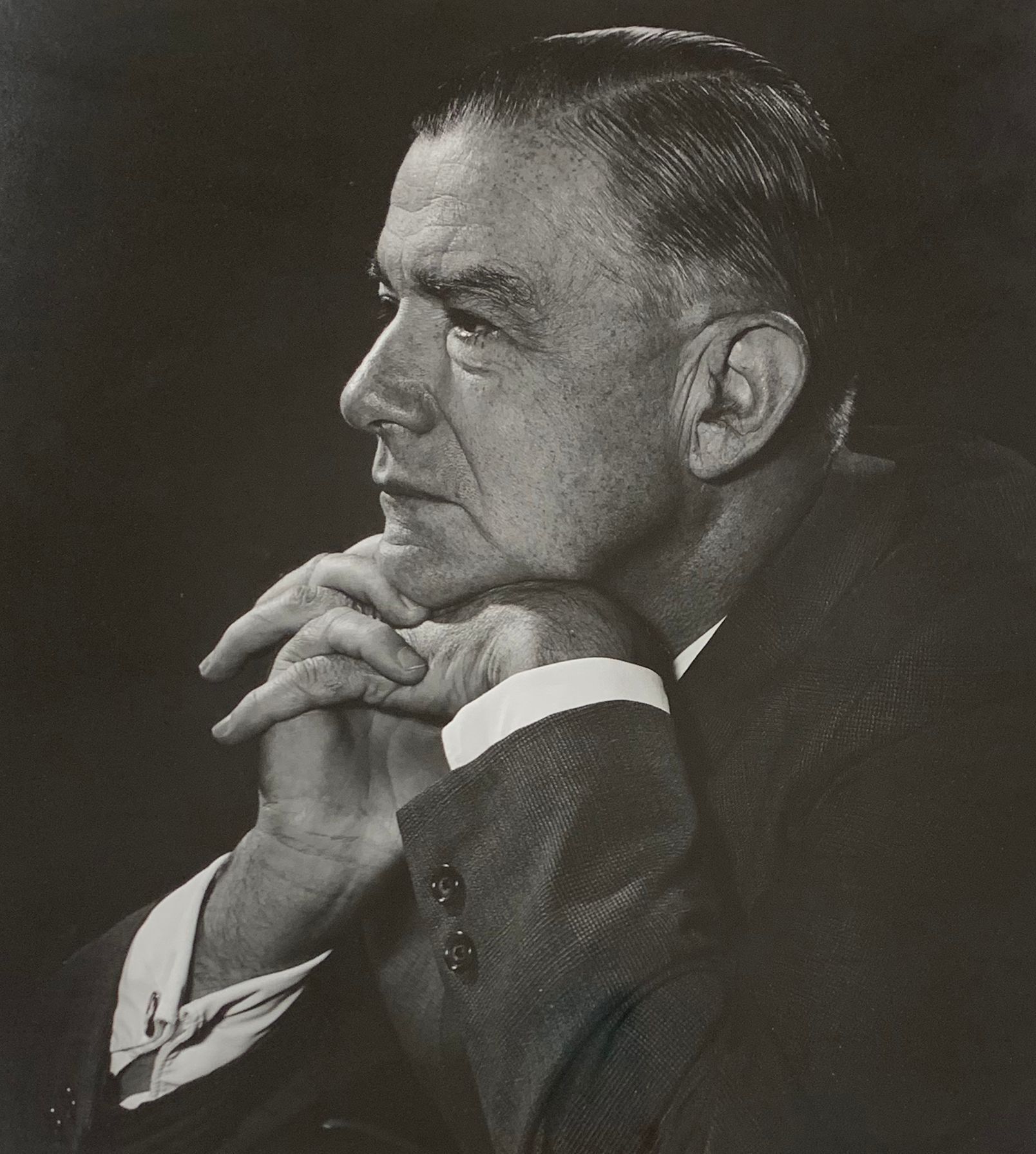 The image depicts a distinguished gentleman, Dr. Paul Brand, pioneer surgeon in profile, dressed in a suit, with a poised and contemplative expression, resting his chin on his hands. His mature features, slicked-back hair, and thoughtful demeanor are emphasized in the black-and-white photograph, highlighting a sense of wisdom and experience.