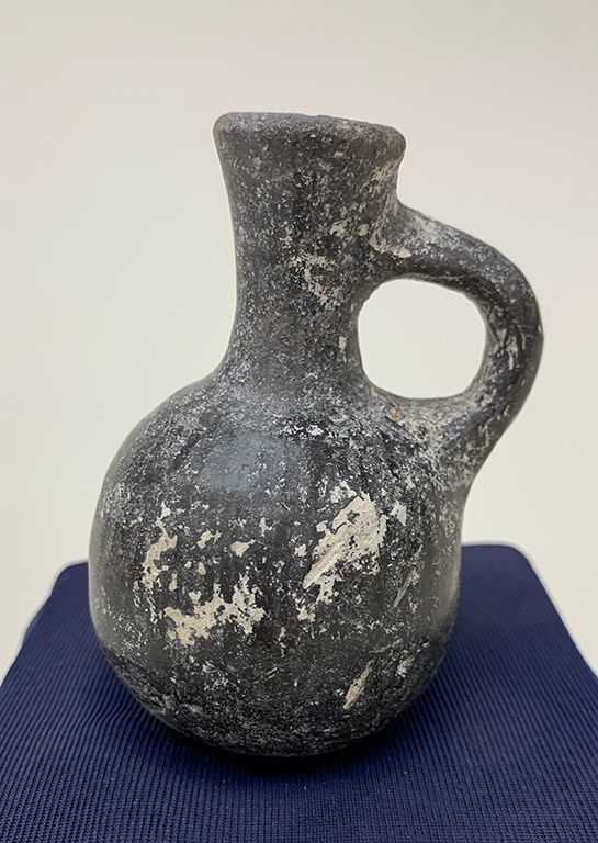 The image features an ancient-looking, small ceramic vessel with a single handle, showcasing a weathered, patterned black surface with white speckles, indicative of its historical wear and aged patina. Its rounded body transitions to a narrow neck, sitting atop a dark blue fabric or mat, highlighting its rustic appearance.