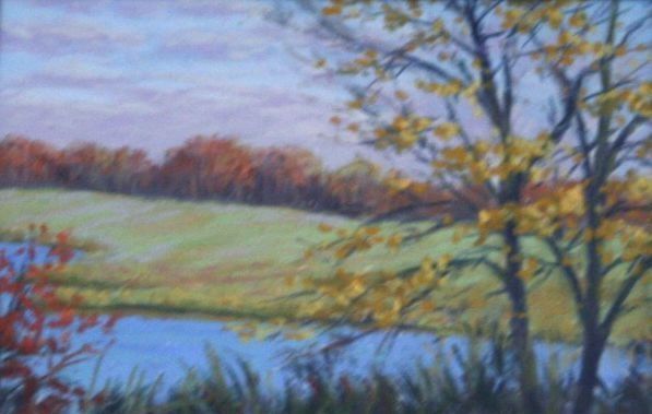 The image depicts a serene autumn landscape featuring a tranquil pond in the foreground with lush grasses and reeds growing along the water's edge. A tree with golden leaves stands prominently on the right, partially obscuring the view of the gently rolling green hills beyond. The background is filled with a line of trees showing vibrant fall color.