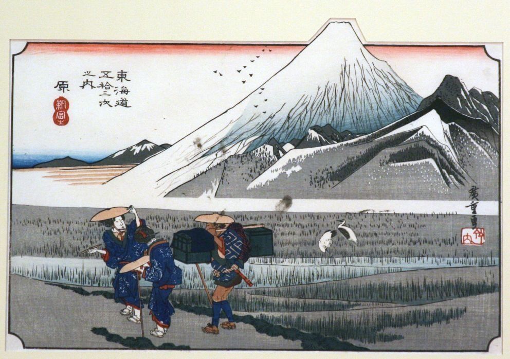 The image depicts a traditional Japanese woodblock print featuring Mount Fuji in the background, with its snow-capped peak rising majestically. In the foreground, two travelers wearing traditional attire and straw hats walk along a path, carrying luggage. Birds are flying in the sky, and two cranes are wading in a marsh.