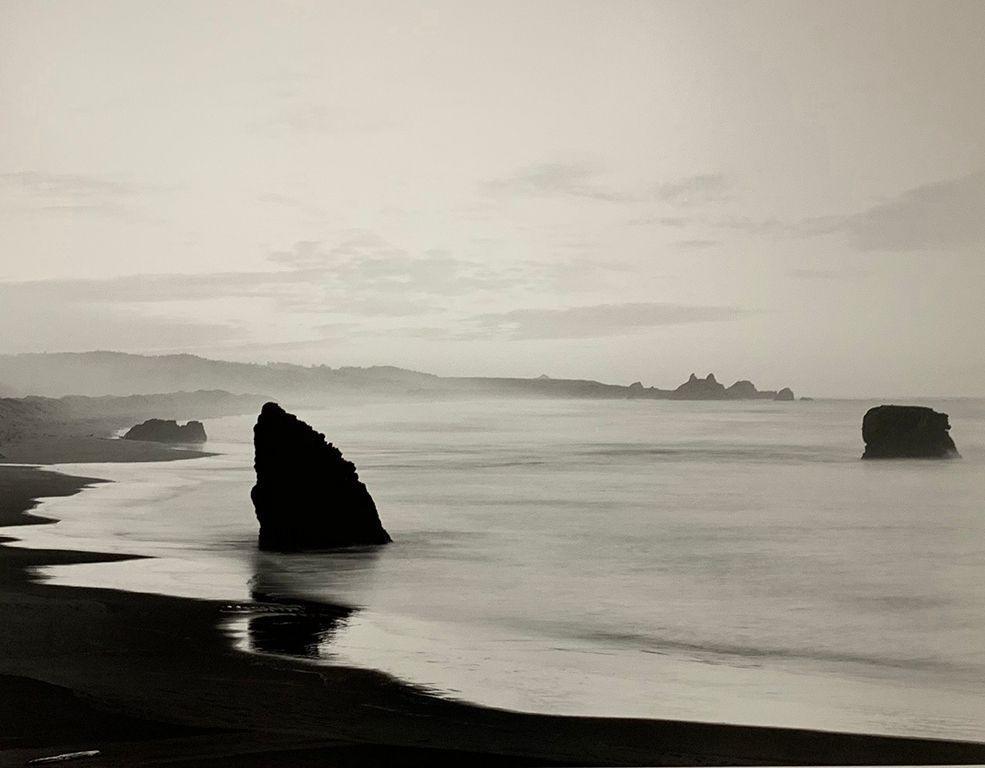 The image depicts a serene seascape in black and white, featuring rugged rocks emerging from the water along a tranquil coastline under an expansive sky with scattered clouds. A peaceful, timeless, and slightly mysterious atmosphere is created that evokes a sense of solitude and reflection, typical of early morning or dusk.