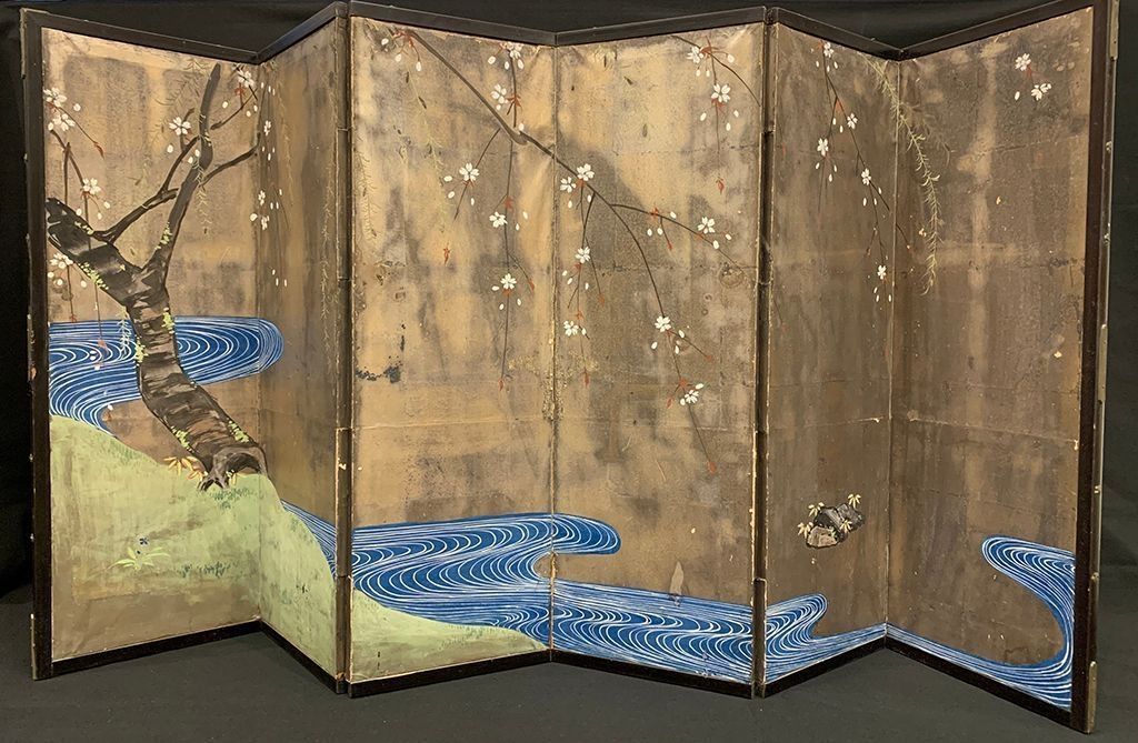 The image depicts a traditional folding screen adorned with a serene scene featuring a winding blue river, a gently leaning tree with delicate branches, and blooming white flowers against a textured, muted background. The vibrant blue of the river creates a striking contrast with the earthy tones, while the blossoms add an ethereal touch to the scene.