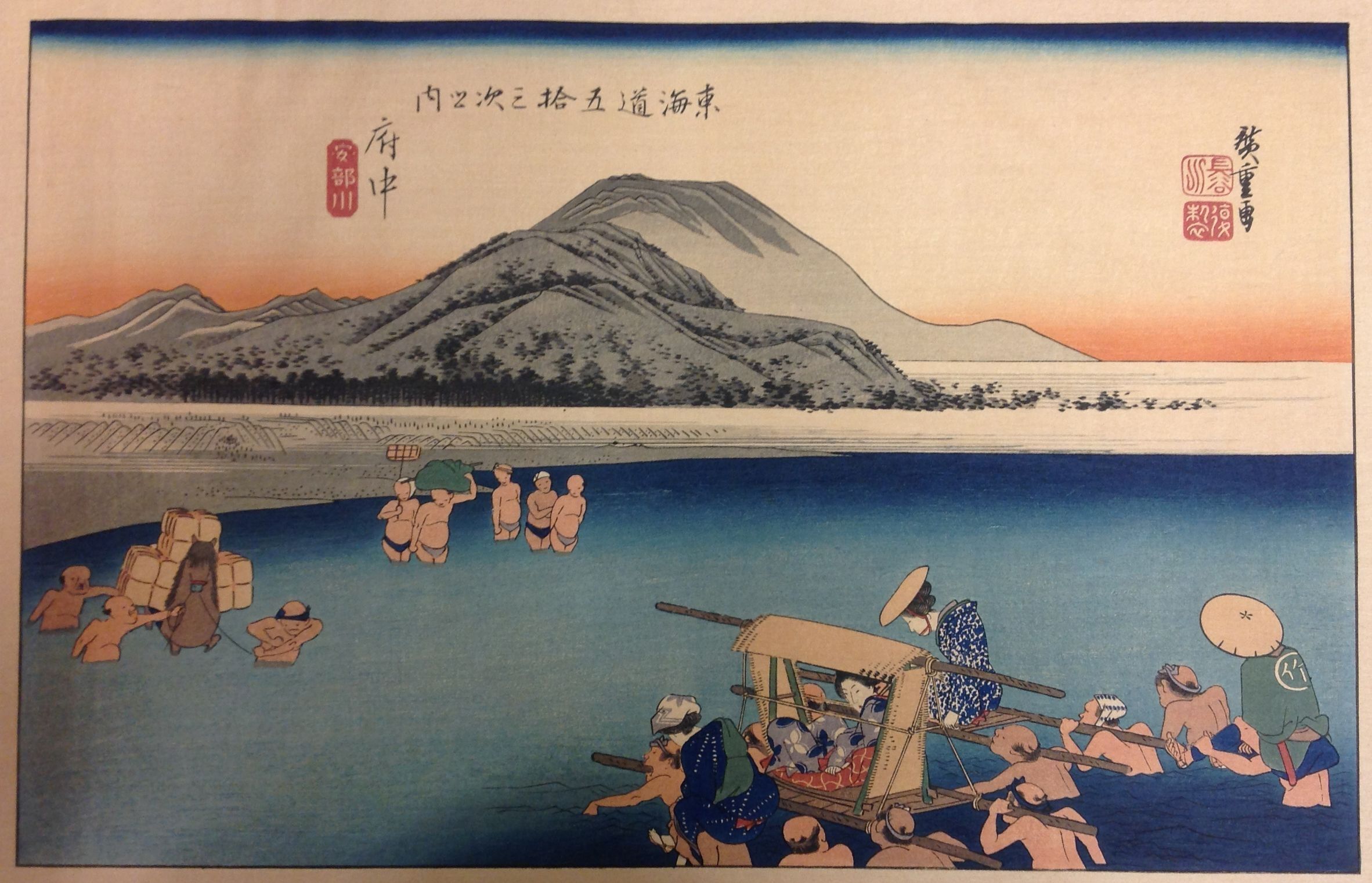 This image is a traditional Japanese woodblock print depicting a scenic river crossing, where several figures are wading through the water, some carrying goods, while others transport passengers in a small boat, set against a backdrop of rolling hills and an expansive sky with a sunset on the horizon.