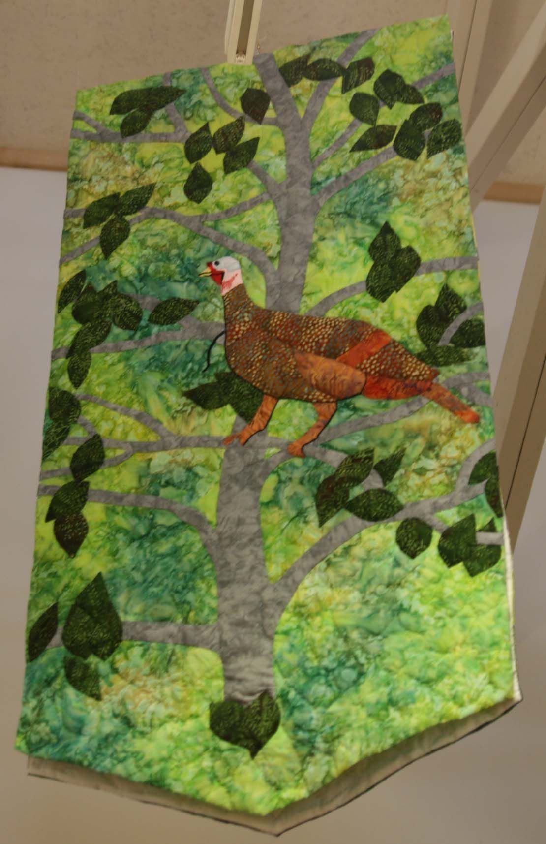A colorful quilted wall hanging depicts a turkey perched on a branch of a tree. The background consists of lush green leaves and a textured trunk, creating a lively forest scene. The turkey, detailed with intricate patterns, stands out against the vibrant tapestry, showcasing a blend of brown, orange, and white feathers.