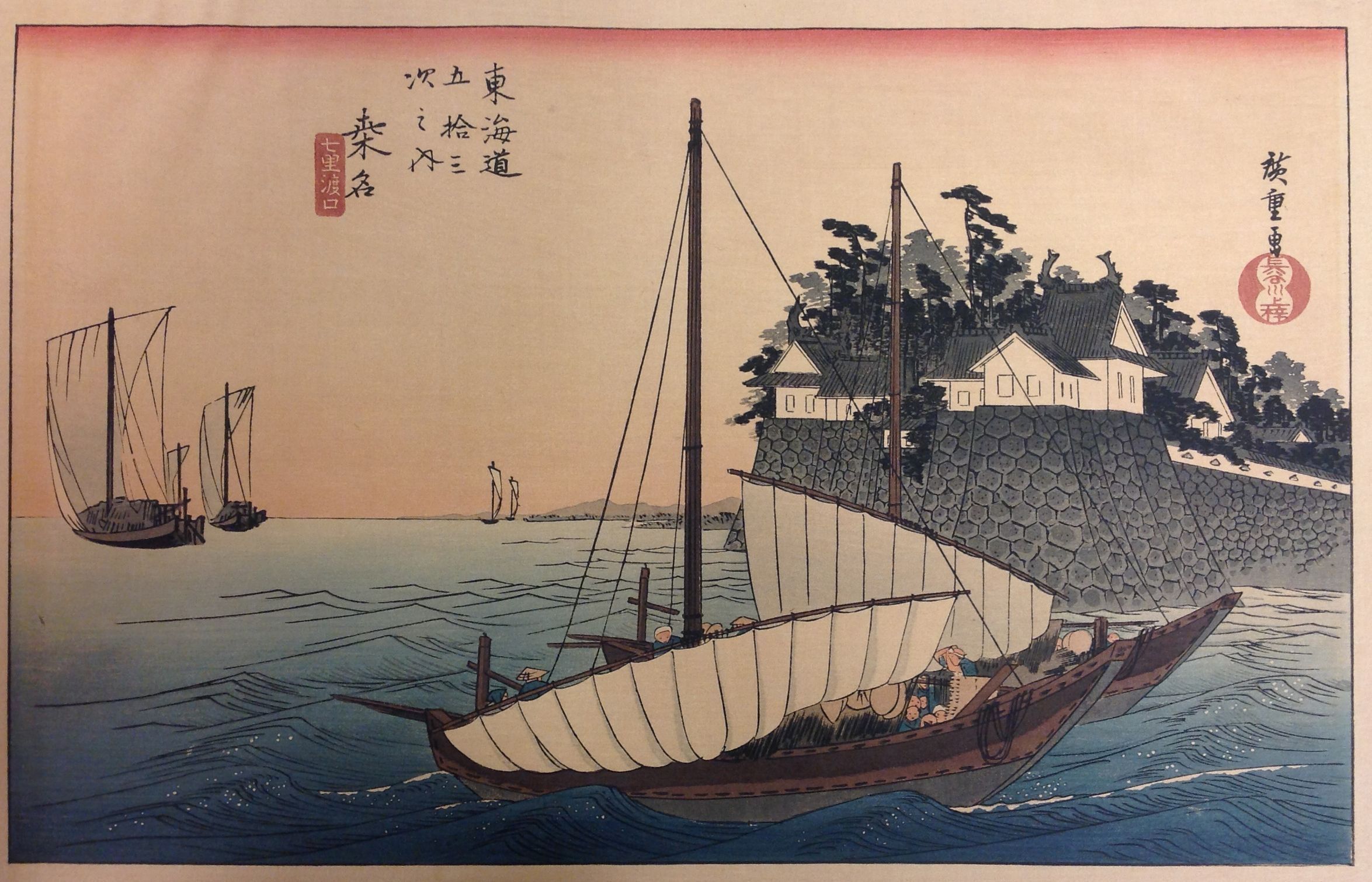 The image depicts a traditional Japanese artwork showcasing sailboats navigating through blue waters near a rocky coastline. The sailboat in the foreground features large white sails and several people visible onboard. Further along the coast, traditional Japanese buildings with sloping roofs are nestled among lush greenery, against a backdrop of mountains.