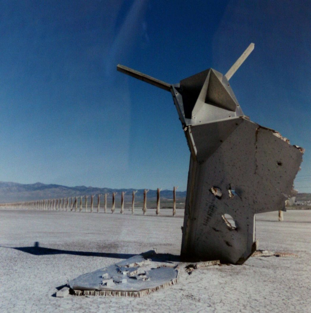 The image depicts a barren landscape featuring a large, partially destroyed, metallic structure with jagged edges, standing prominently on a cracked, dried surface under a clear blue sky. In the background, a long line of evenly spaced wooden posts stretches across the vast, arid expanse towards a distant mountain range.