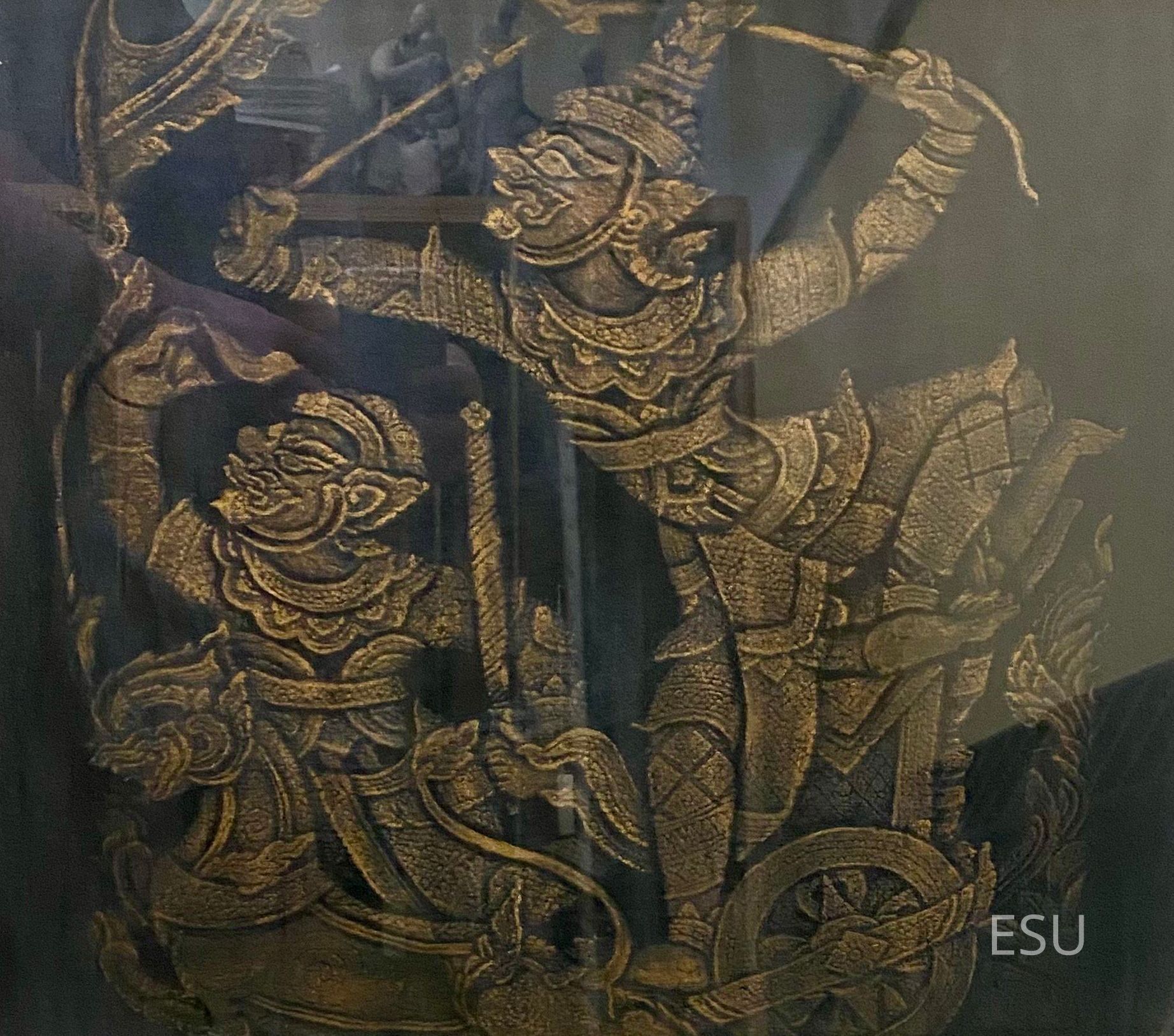 "Thailand Gods" by Herbert Seiler: The image shows a detailed, ornate carving depicting two mythological figures in traditional attire, engaged in dynamic movements with flowing garments and intricate accessories, one of them holding a bow and readying an arrow, while the other is poised beside an elaborate chariot wheel, all rendered in a striking gold tone that enhances the intricacies and textures, creating a sense of movement and energy that captures a scene rich with cultural symbolism and storytelling, set against a dark background that adds contrast and depth to the artwork, highlighting