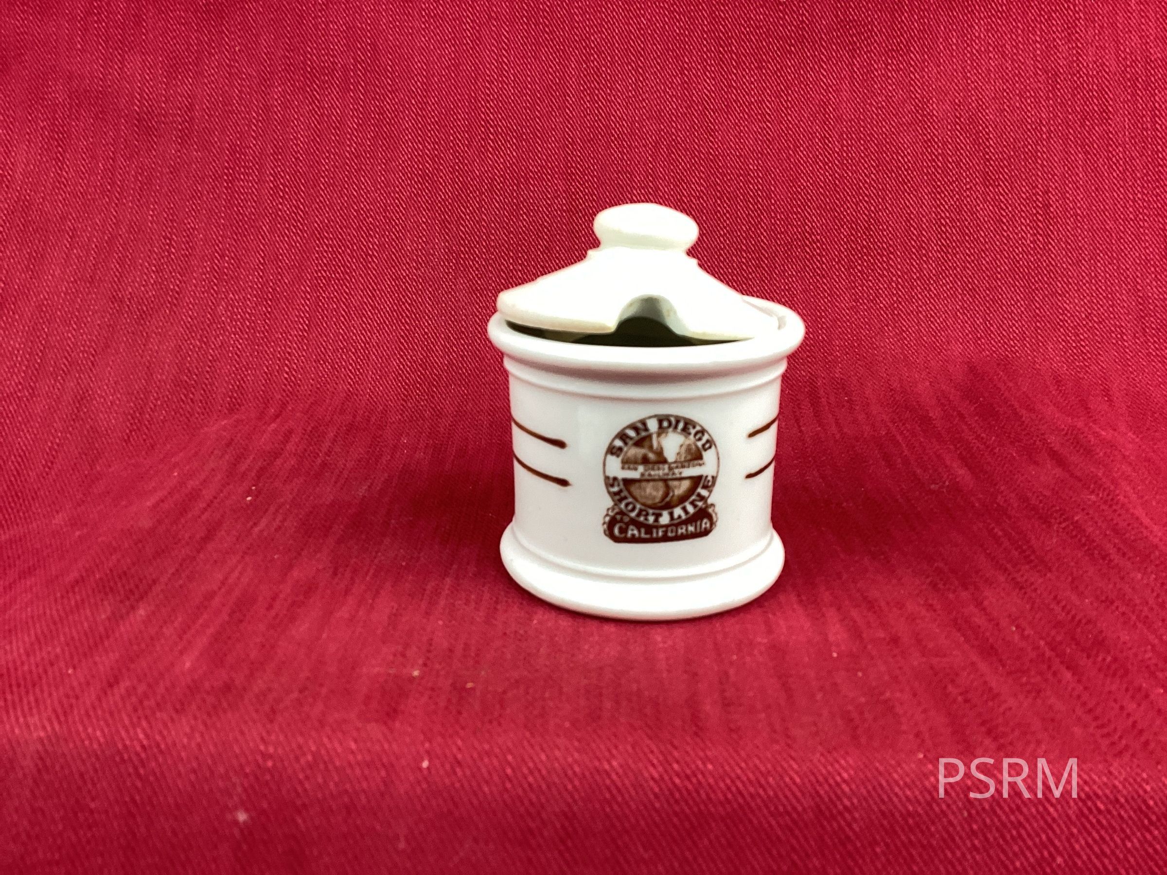 The image depicts a small, white ceramic container with a lid, adorned with the text "San Diego Short Line California" and a decorative emblem on its front. The container is set against a rich, red textured fabric background, which adds a warm contrast to the white and brown hues of the ceramic. The overall presentation of the object and its setting suggests a quaint, vintage feel, possibly invoking a sense of nostalgia or connection to a specific locale. The lid of the container appears slightly ajar, adding