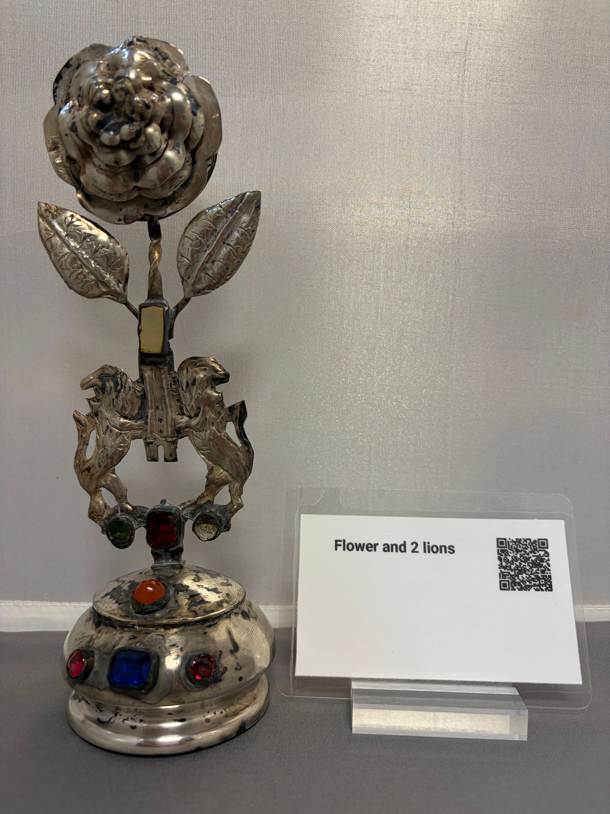Silver spice box used for Saturday night Havdalah: The image features an ornate silver object, consisting of a large, intricately detailed flower with two leaves, stemming from the base where two lions are depicted. The base of the object is adorned with colored gemstones in red, blue, orange, and green, creating a vibrant contrast against the metallic finish. To the right, a small placard labeled "Flower and 2 lions" is present with a QR code printed alongside. The whole arrangement rests on a gray surface set against a light-colored background,