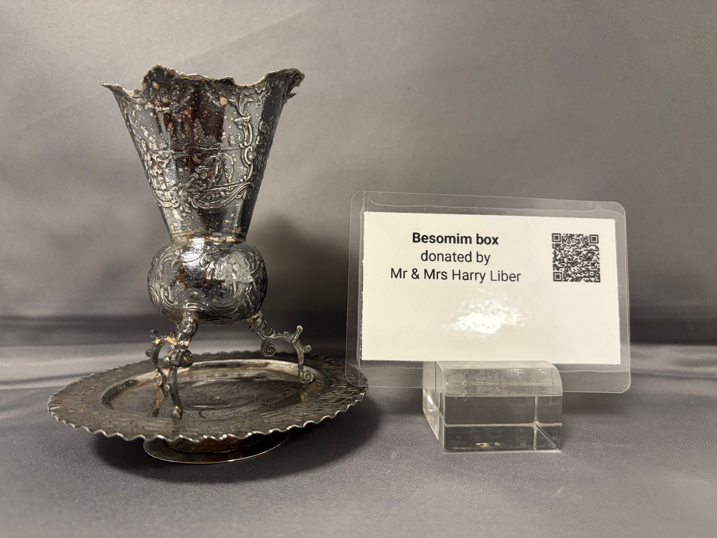 The image shows an ornately designed Besomim Box from Israel, intricately crafted with embellished details, resting on a small platter within a display case. Adjacent to it is a label indicating that this item was gifted by Mr. and Mrs. Harry Liber for the confirmation of Susan Beth Liber. The Besomim Box, used in Jewish rituals, appears to be made of metal with a decorative pattern that highlights its cultural and historical significance.