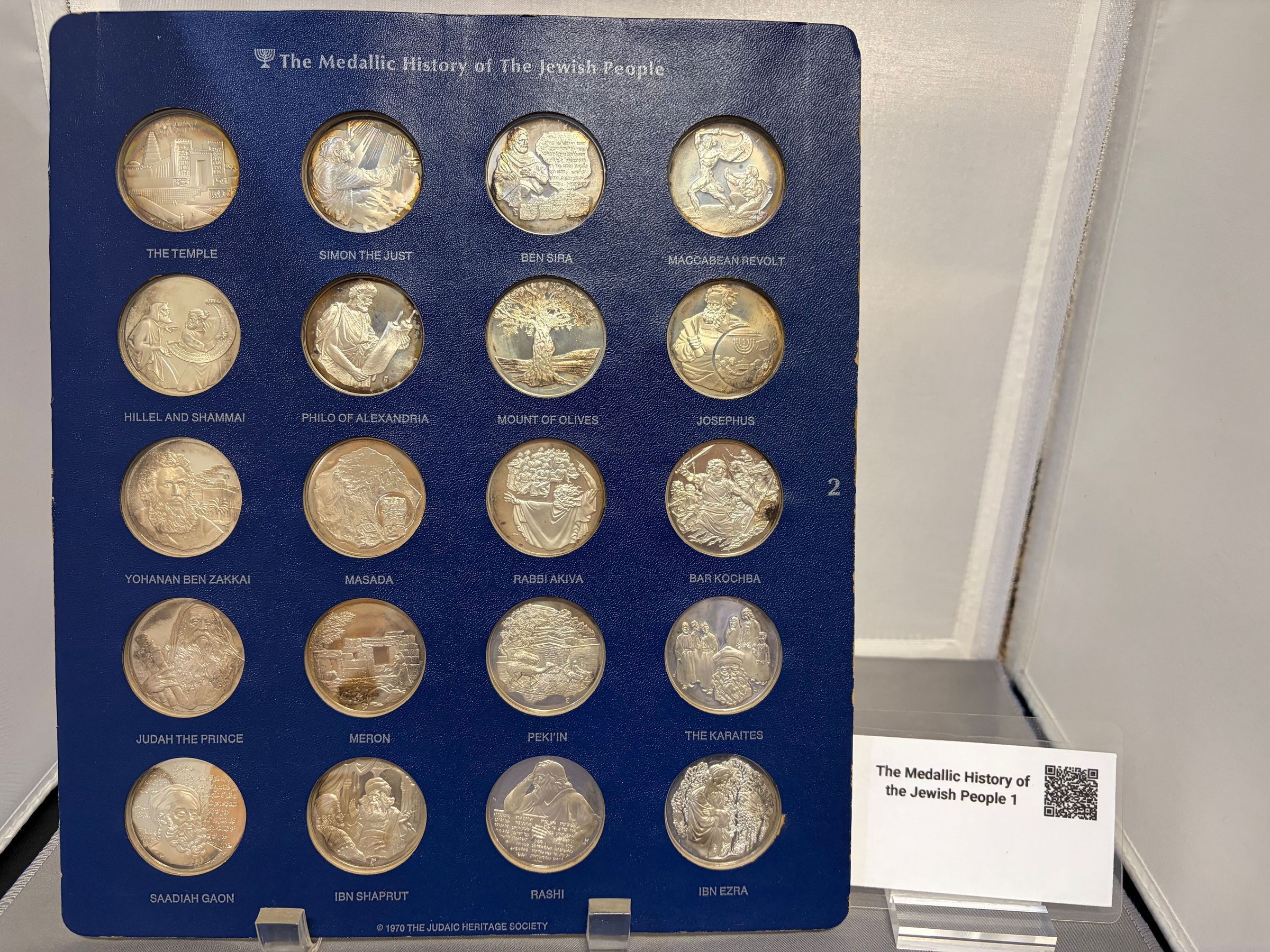 The Medallic History of the Jewish People (2): The image displays a collection of commemorative coins encased in a blue display holder titled "The Medallic History of The Jewish People," featuring inscriptions and reliefs that represent various significant historical and cultural figures or events such as "The Temple," "Simon the Just," "Ben Sira," "Maccabean Revolt," "Hillel and Shammai," "Philo of Alexandria," "Mount of Olives," "Josephus," "Yohanan Ben Zakkai,"