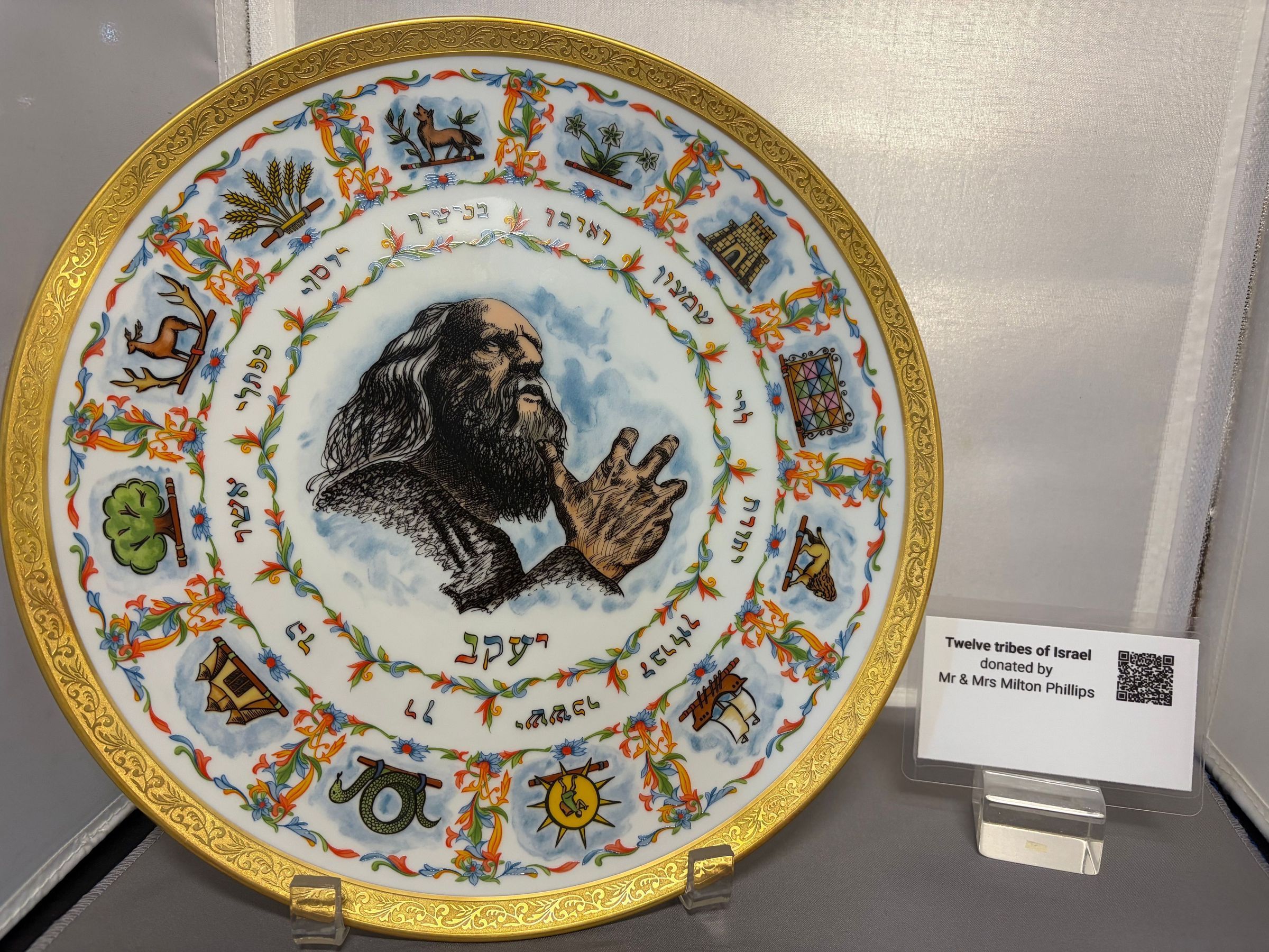 Twelve Tribes of Israel: The image displays an intricately designed decorative plate featuring an illustration of a figure with a beard at its center, surrounded by colorful motifs and symbols representing the Twelve Tribes of Israel, with Hebrew inscriptions around the edge; the plate, set on a transparent stand, is accompanied by a label that reads: "TWELVE TRIBES OF ISRAEL, First Edition by Laszlo Ispanky, Gift of Mr. and Mrs. Milton Phillips in honor of the Confirmation of Larry Phillips."