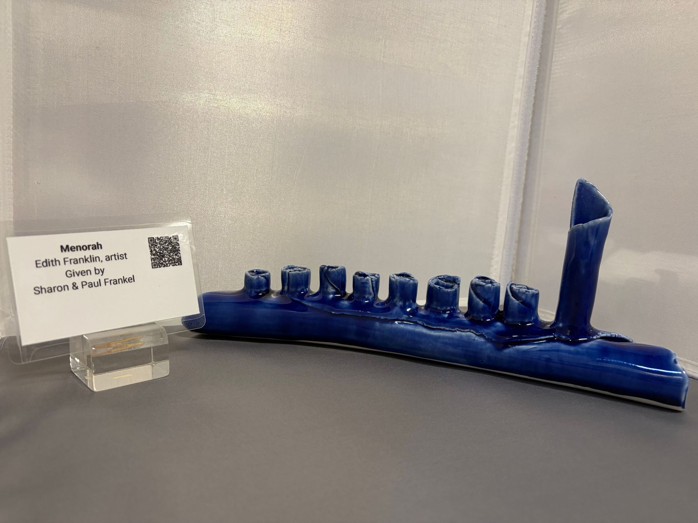 Menorah, Edith Franklin, Artist: The image depicts a blue ceramic menorah with nine candle holders arranged in a horizontal line, with the tallest holder on the far right side. It is displayed against a light background on a gray surface. Next to the menorah is a small sign mounted on a clear stand, providing information about the piece. The sign indicates that the menorah was created by the artist Edith Franklin and was given by Sharon and Paul Frankel.
