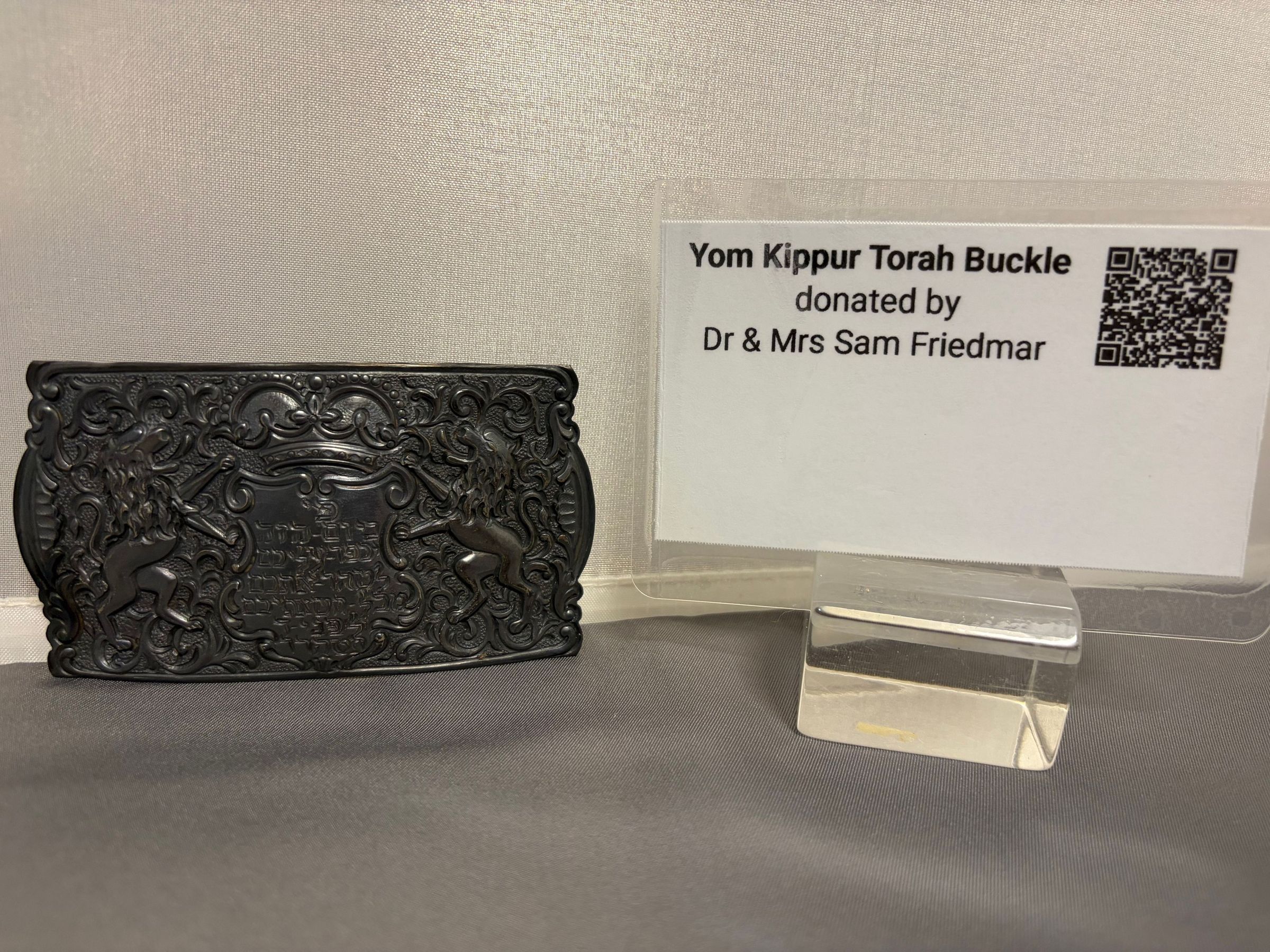 Yom Kippur Torah Buckle: The image features a Yom Kippur Torah buckle, intricately designed with ornate, symmetrical patterns and figures, possibly depicting lions, set against a background that highlights their detailed craftsmanship; alongside the buckle, there is a clear display stand holding a sign that reads "Yom Kippur Torah Buckle donated by Dr & Mrs Sam Friedmar" with a QR code on the right side, all positioned on a textured fabric surface that complements the dark metal of the buckle and the contemporary simplicity of the object.