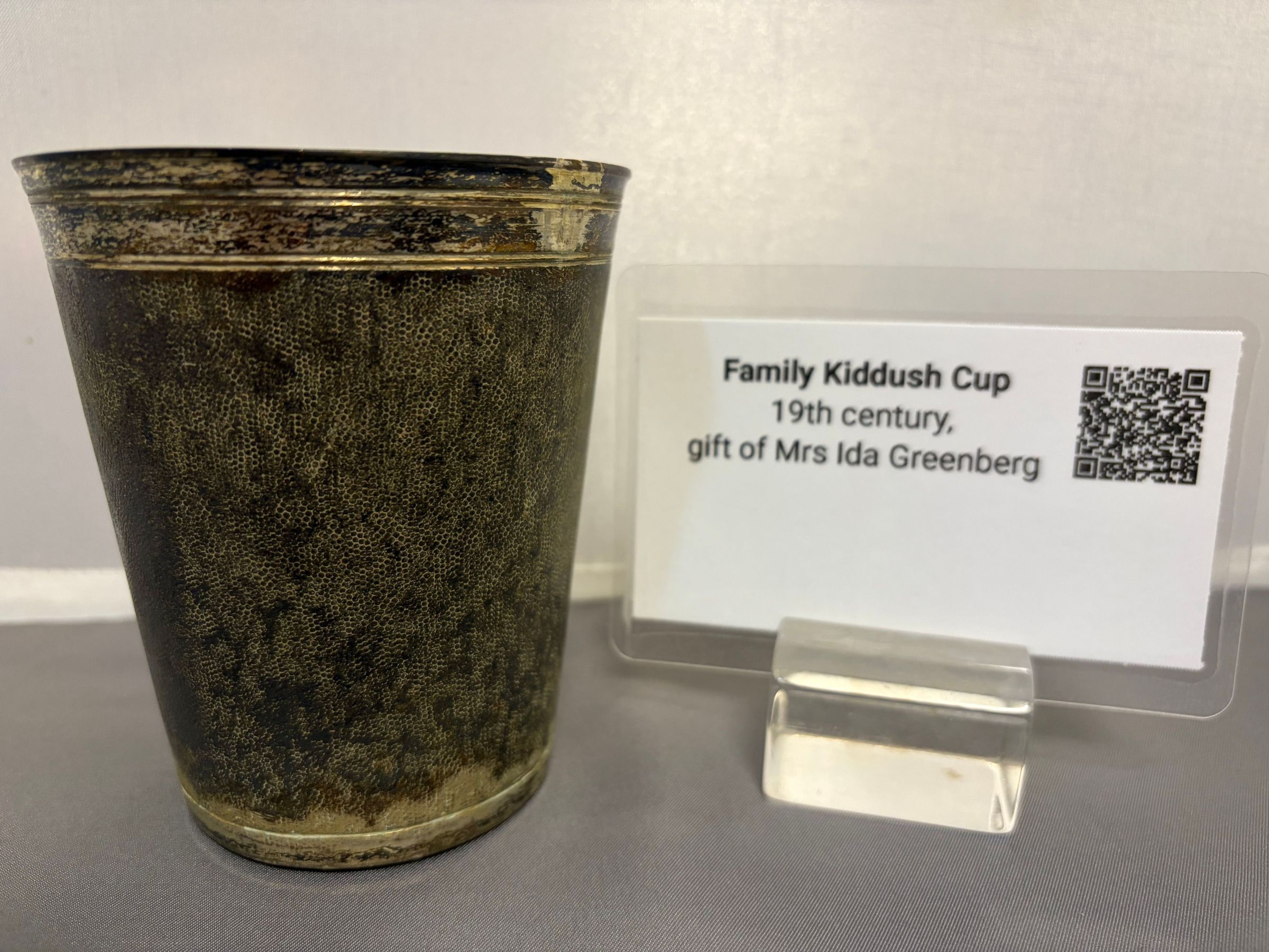 Family Kiddush Cup 19th century: The image depicts a dark, textured family Kiddush cup, which is presented alongside a small informational plaque. This plaque, held in a clear stand, identifies the item as a 19th-century Kiddush cup and notes that it was a gift from Mrs. Ida Greenberg. A QR code is also included on the plaque, suggesting digital access to further information. The scene is set against a plain, light-colored backdrop, emphasizing the cup's historical and cultural value while inviting viewers to appreciate its significance