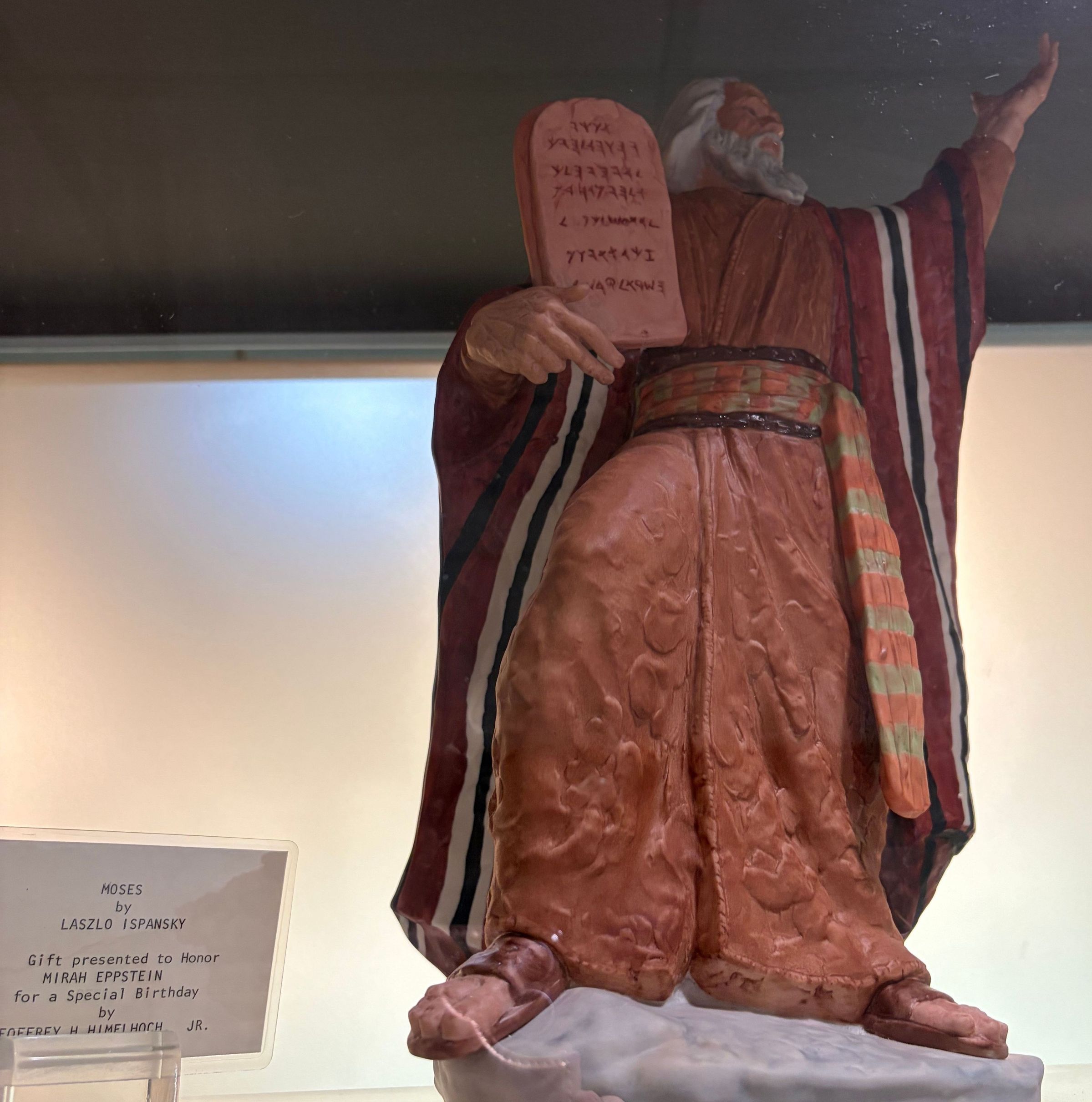 Moses located behind the Library Circulation desk: The image depicts a detailed figurine of Moses holding a tablet with inscriptions, representing the Ten Commandments, while gesturing with his other hand. The figure wears a long robe with a striped pattern and a colorful sash, evoking traditional biblical attire. The figurine is mounted on a base, and nearby, a plaque reads, “MOSES by LASZLO ISPANSKY," indicating it was a gift to honor Mirah Eppstein for a special birthday from Geoffrey H. Himelhoch, Jr.