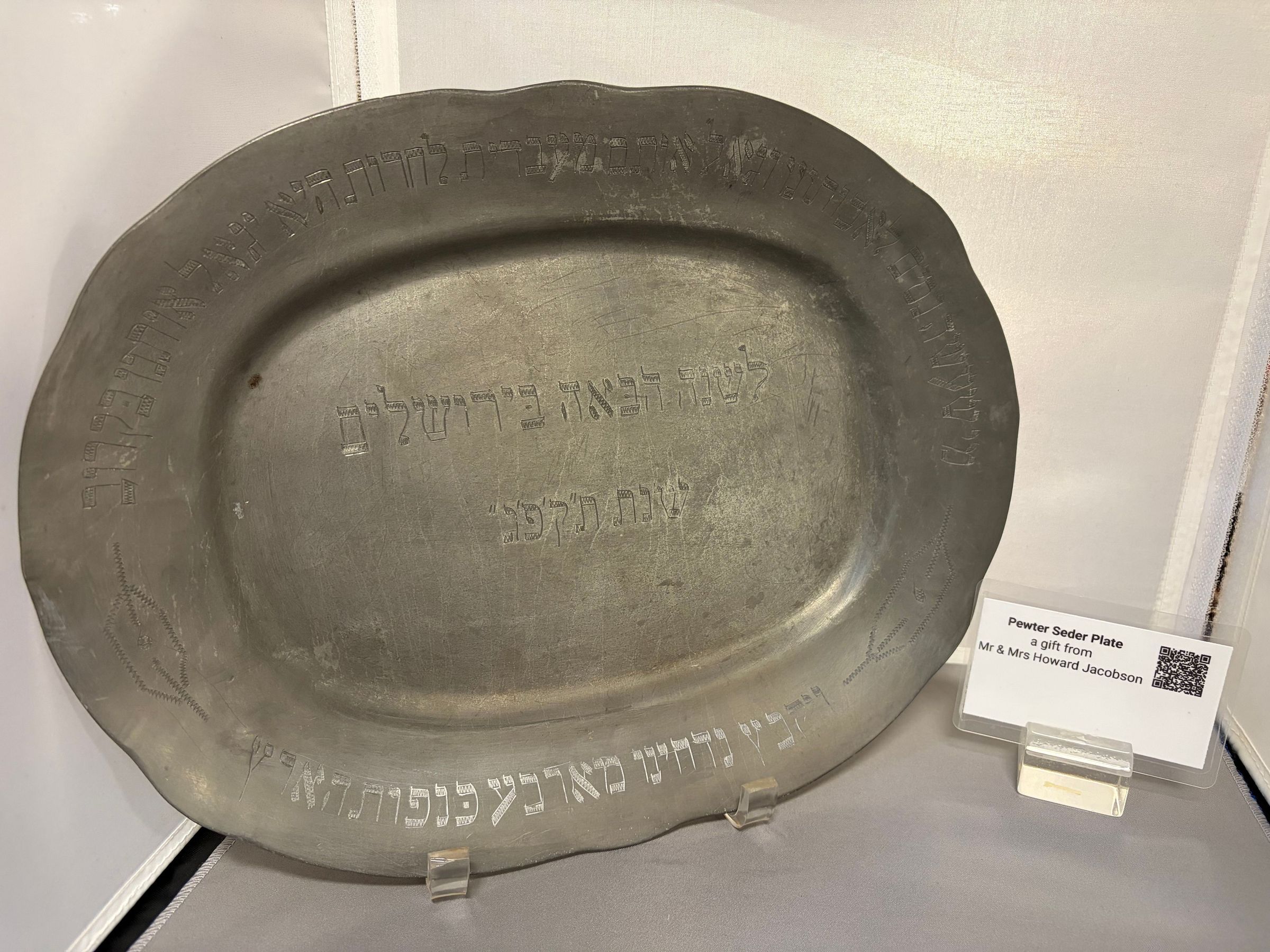Pewter Seder plate: The image shows an early 19th-century pewter Seder plate, which is displayed behind a protective glass case. The plate features embossed Hebrew text around its rim, reflecting its cultural and religious significance, and it is noted as a gift from Mr. and Mrs. Howard Jacobson, presented in memoriam of Mrs. Cora Hahn Jacobson, as indicated by a small label beside the plate.