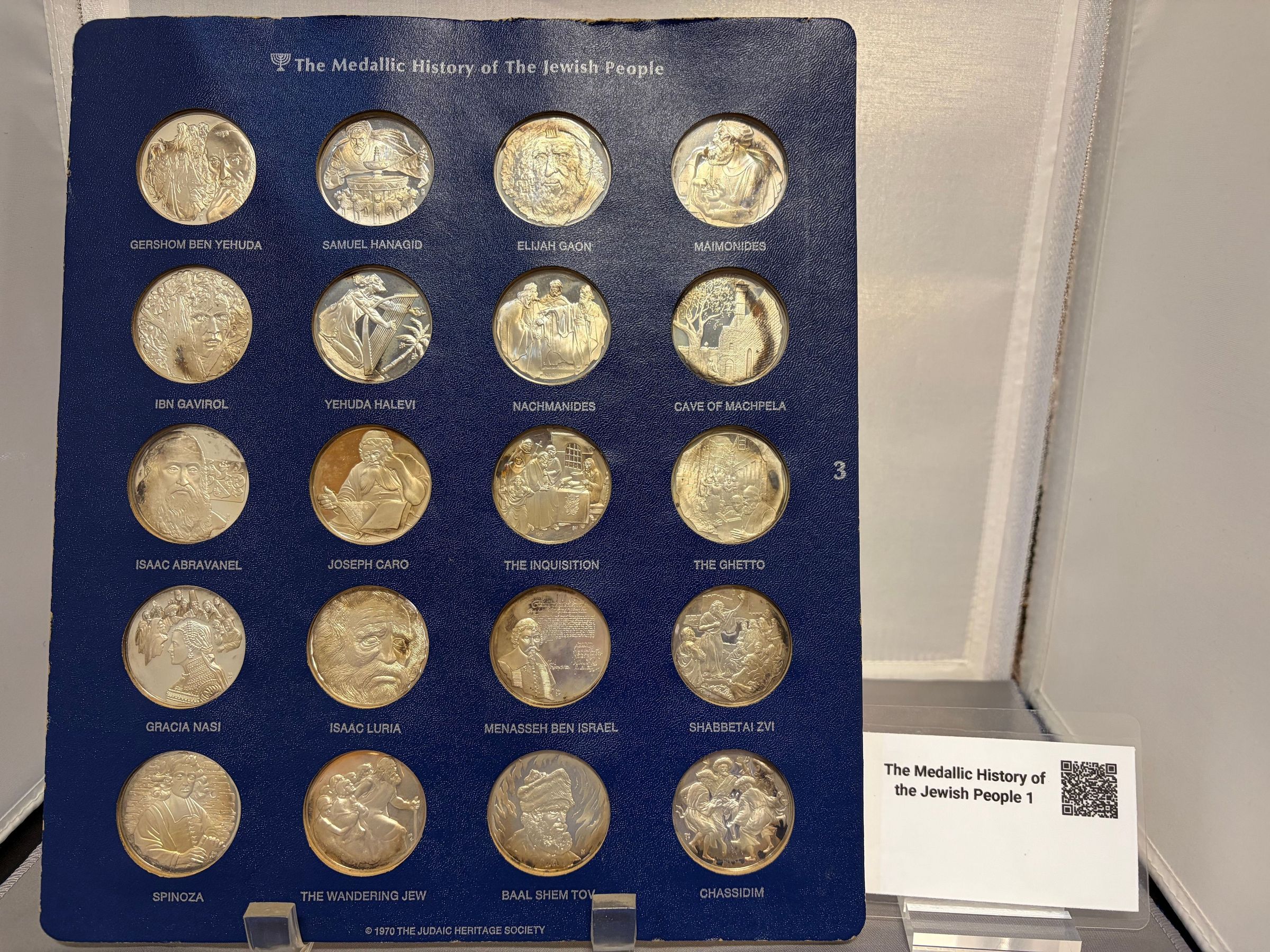 The Medallic History of the Jewish People (3): The image displays a blue panel titled "The Medallic History of The Jewish People" containing twenty silver medals, each featuring a different design and name beneath them. These include notable figures and significant themes such as Gershom Ben Yehuda, Samuel Hanagid, Elijah Gaon, Maimonides, and others like The Inquisition and The Ghetto. Each medal is carefully placed within individual compartments, and a small sign beside the panel reads "The Medallic History of the Jewish People 1