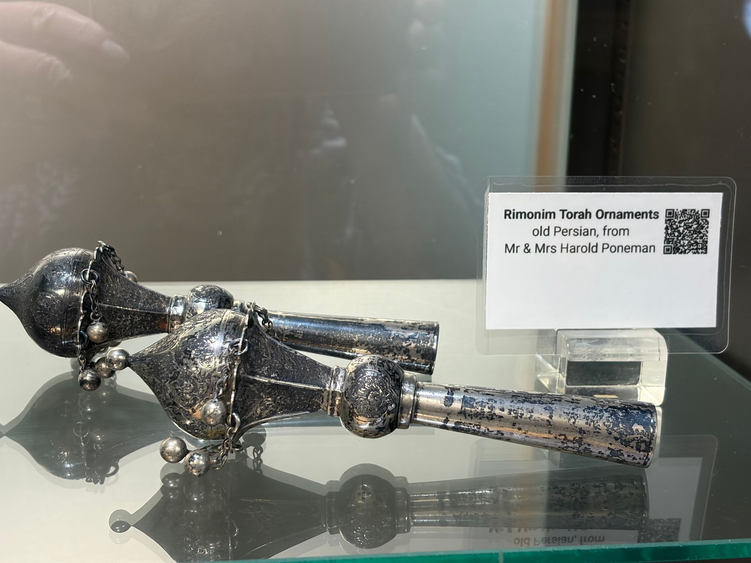 The image shows two ornate, antique silver rimonim, which are decorative finials used on Torah scrolls, featuring intricate designs and small dangling bells, displayed on a glass surface with a label beside them reading, "Rimonim Torah Ornaments, old Persian, from Mr & Mrs Harold Poneman," indicating their cultural and historical significance.