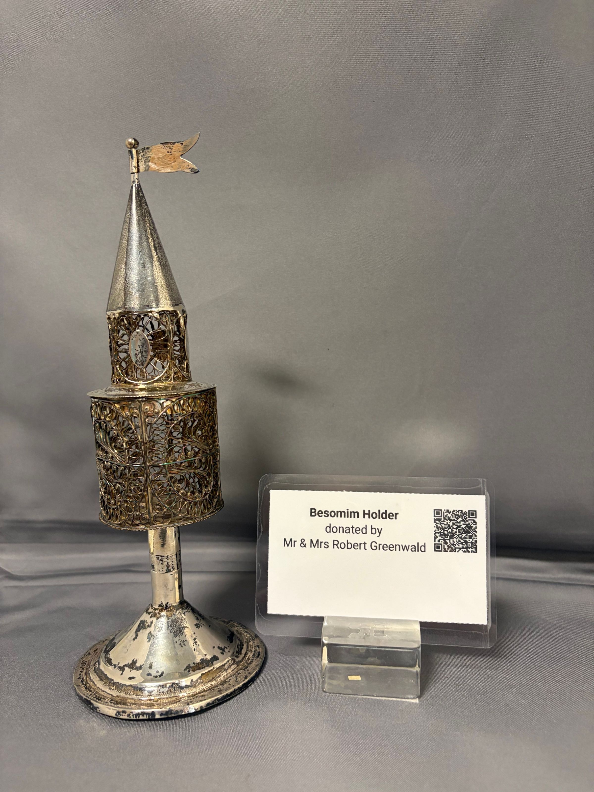The image shows an ornate, tower-shaped Besomim holder with intricate silver filigree work and a conical top adorned with a small flag, placed against a plain background. It stands on a circular base that displays signs of aging with a patina, suggesting it is an antique or vintage piece. Next to the holder is a clear plastic stand holding a card that reads, "Besomim Holder donated by Mr & Mrs Robert Greenwald," along with a QR code. The setup appears to