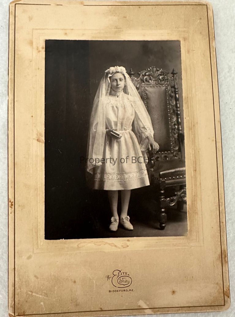 An albumin print on 6 x 9 mat taken at  Elite Studio in white dress and veil with a rosary and book in her hand.