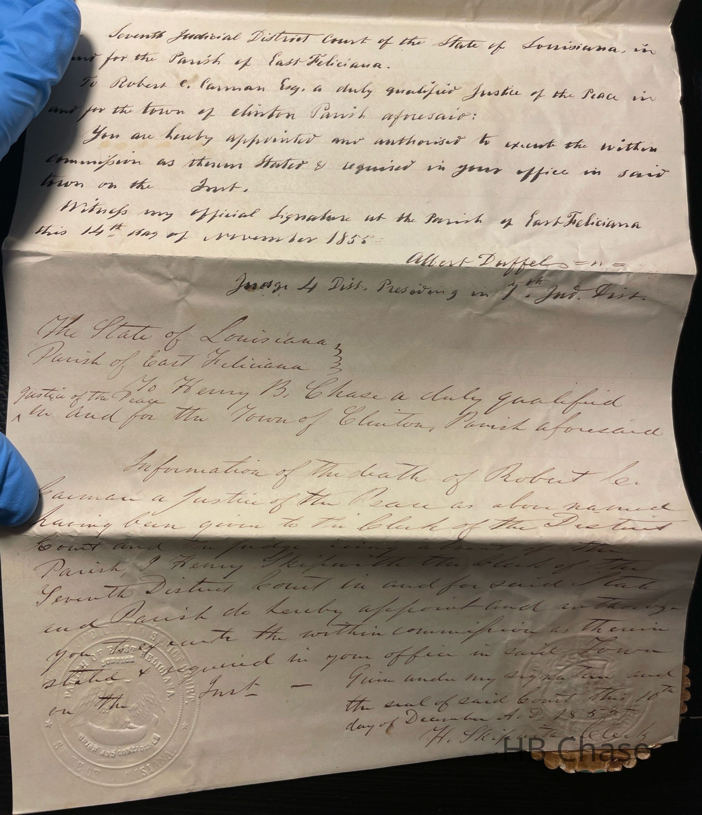 The image shows an old document, written in cursive, which appears to be an official appointment or legal notice. The text is handwritten on yellowed paper, including signatures, and bears an embossed seal. A gloved hand is holding the paper at one corner, indicating careful handling, likely due to its historical significance. The document is titled with references to the State of Louisiana and East Feliciana Parish, mentioning the appointment of a justice of the peace in 1855. It is signed by