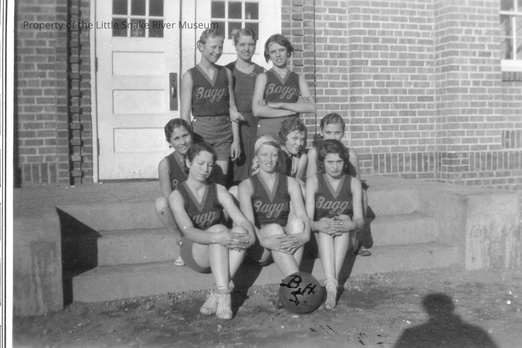 In a black and white photograph, a group of young women, presumably a sports team, poses on the steps in front of a building made of brick, wearing matching sleeveless jerseys with "Baggs" written on them, suggesting they might be representing a school or community team; they are arranged in three rows, with some sitting on the steps and