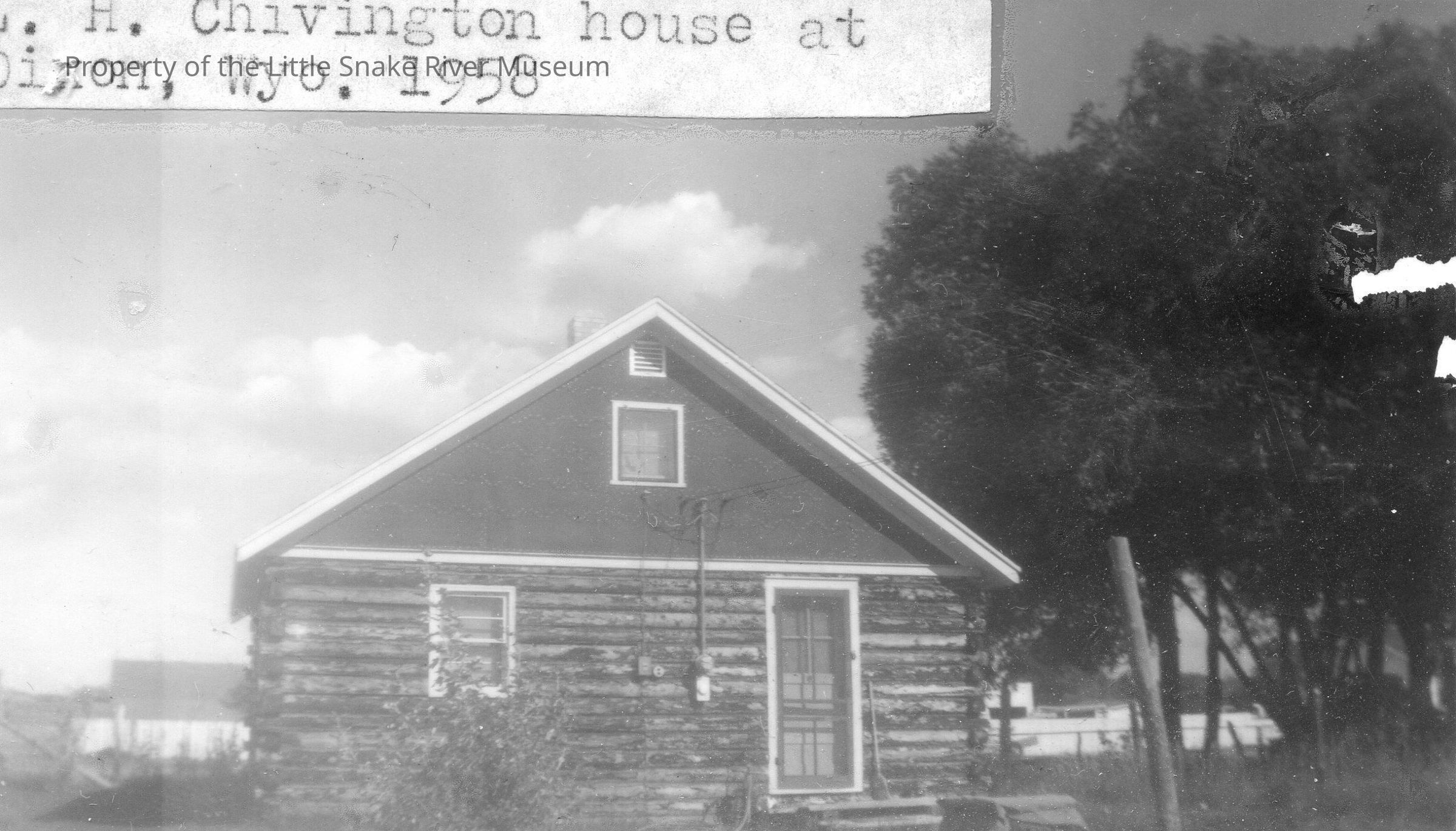 The black and white image features a small log cabin with a steep gabled roof located in a rural setting, surrounded by tall trees and a partly cloudy sky; the house, identified as the H. Chivington house, is positioned in Dixon, Wyoming, as noted by the caption from 1958; a visible electric meter box and a partially obscured wooden fence are also present in the photograph.